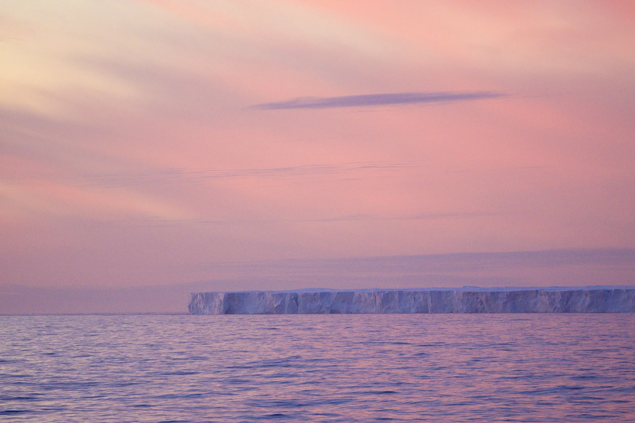 A pink and purple sunset over the ocean with a large iceberg floating in the water.