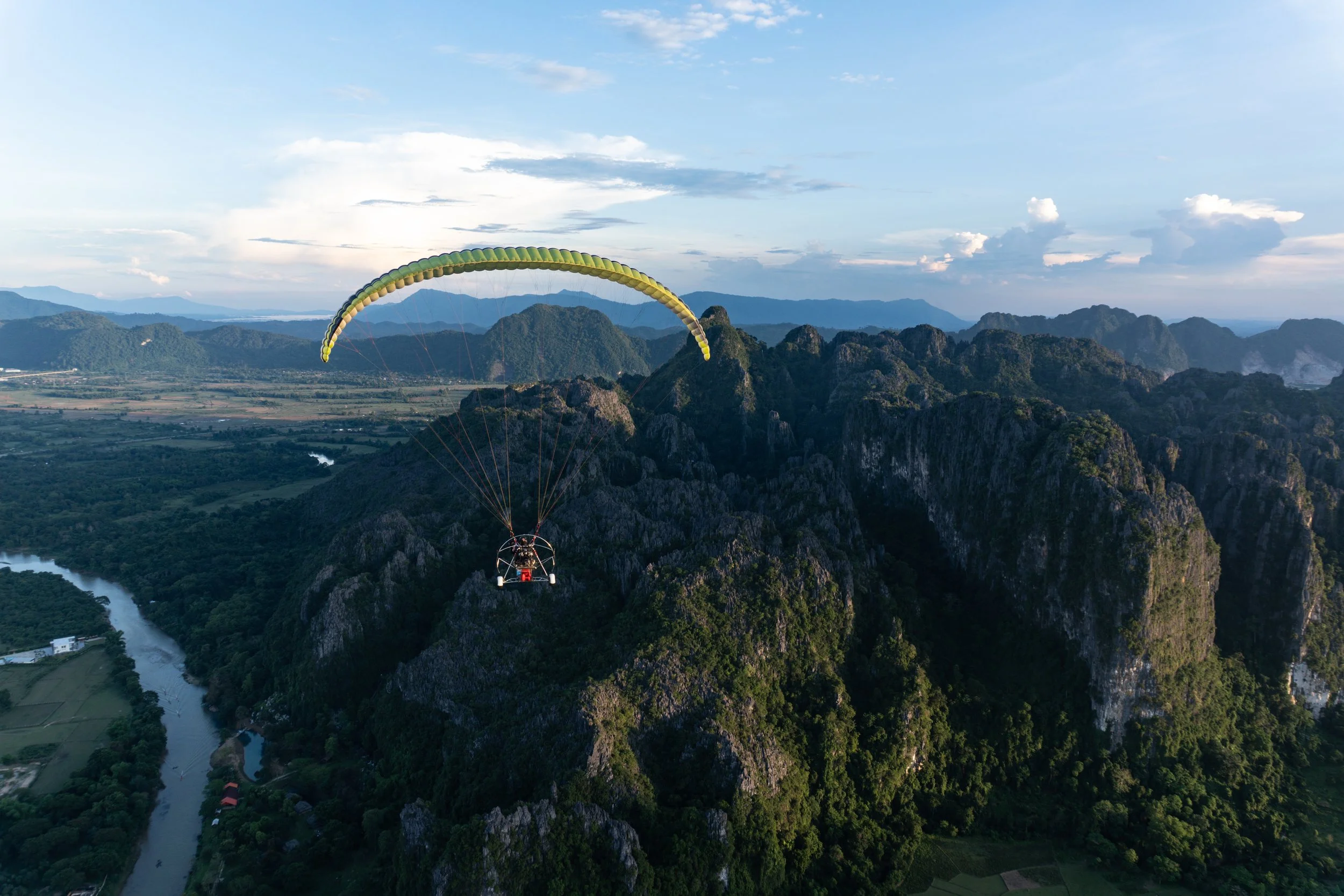 A person paramotoring over lush green mountains and a winding river, with a blue sky and scattered clouds in the background.