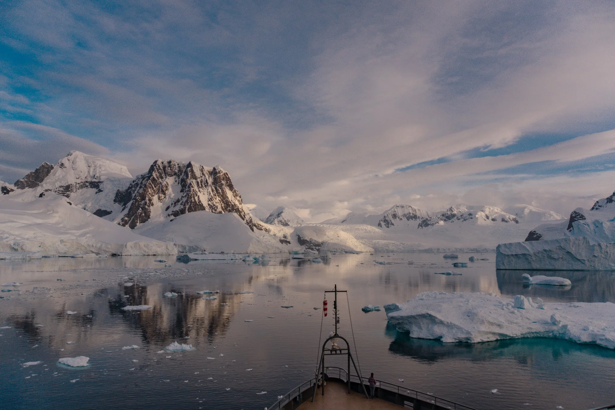 View from a ship's deck of icy Antarctic landscape with snow-covered mountains, floating icebergs, and calm water under a partly cloudy sky.