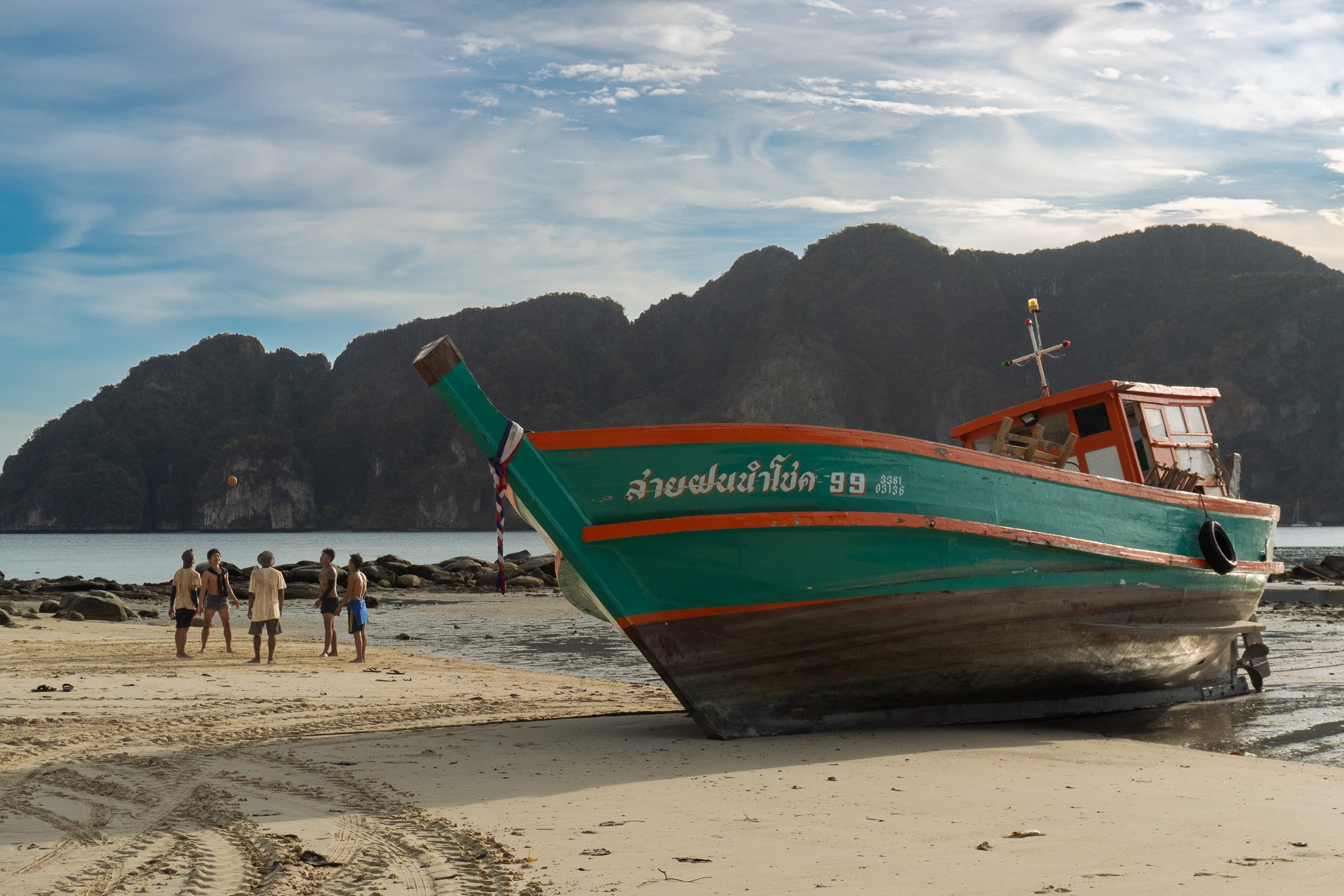 A large boat on a sandy beach with five people standing nearby, with mountains in the background and a partly cloudy sky.