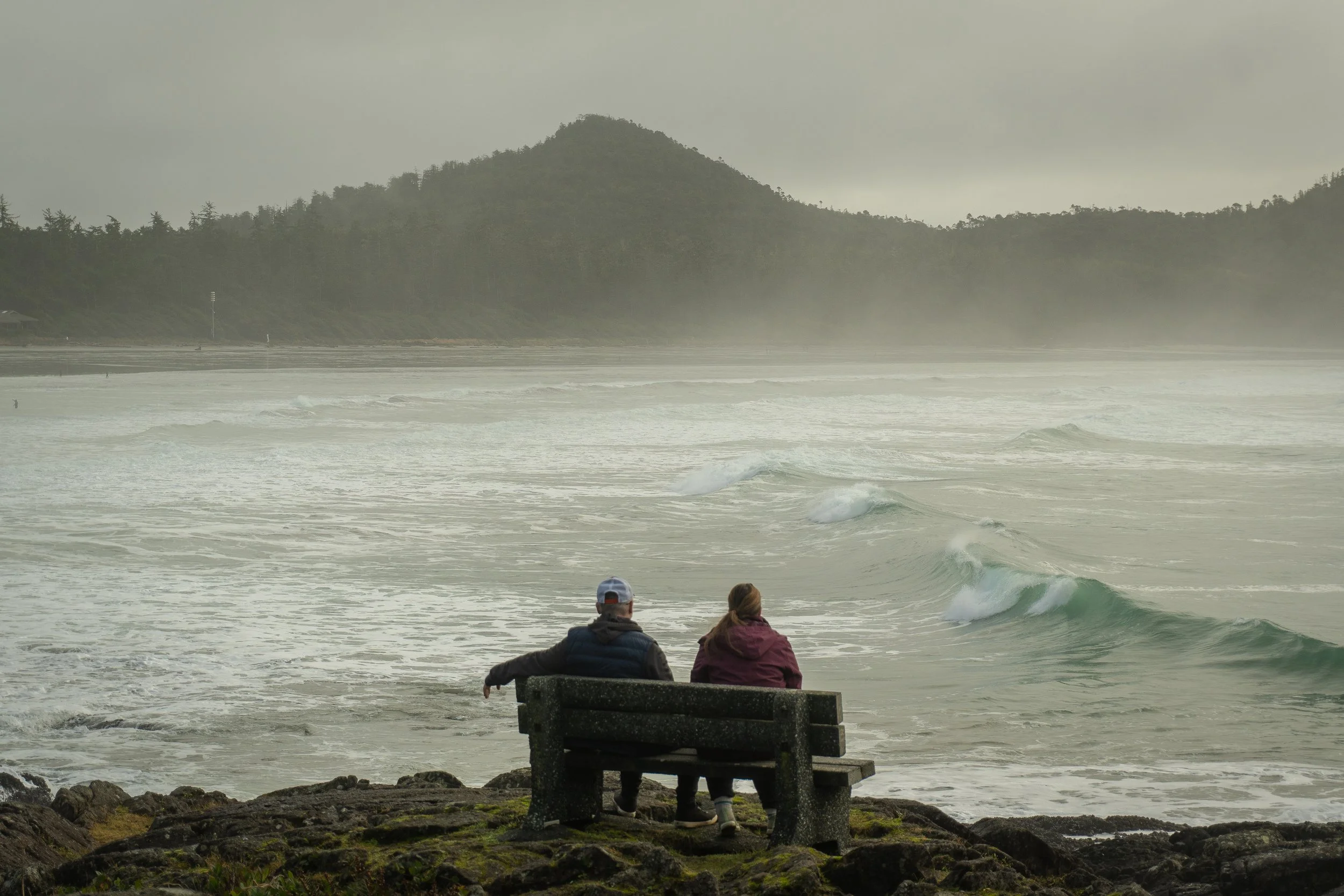 A couple sitting on a bench near the rocky shore, watching the ocean waves on a foggy day with hills in the background.