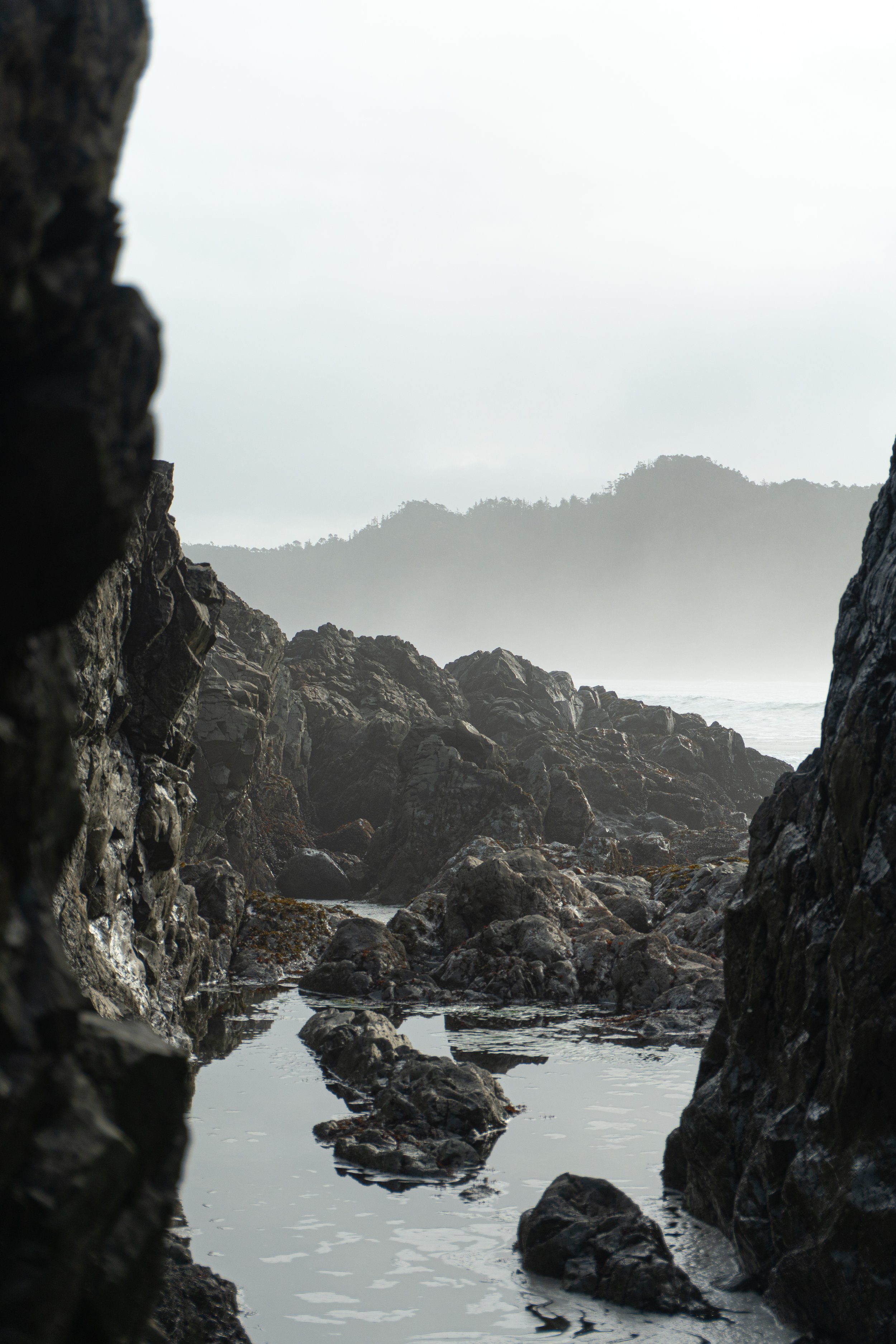 View through a rocky crevice to a misty seascape with large rocks and distant hills.