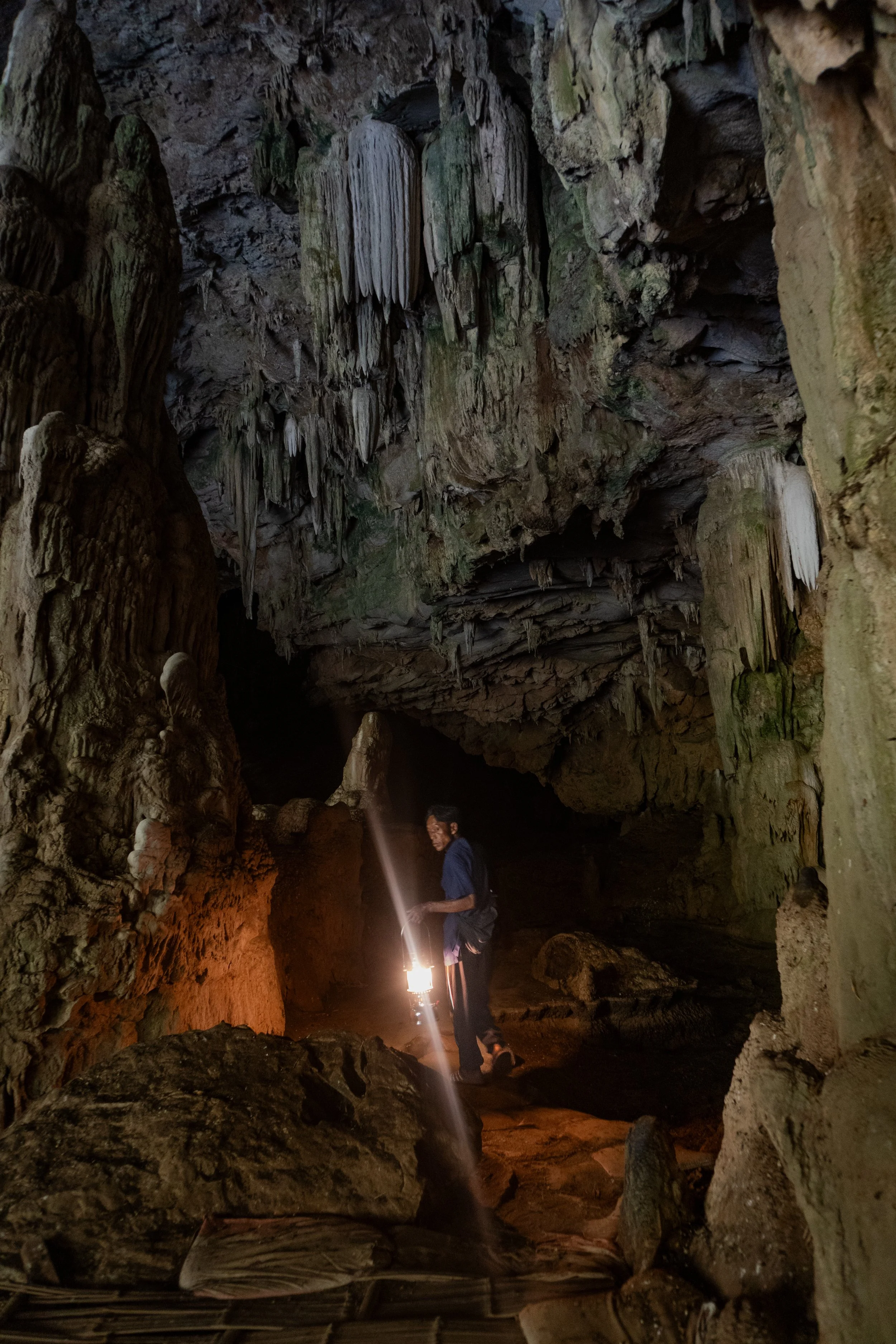 A person exploring a dark cave with stalactites hanging from the ceiling, holding a lantern which illuminates the cave walls.
