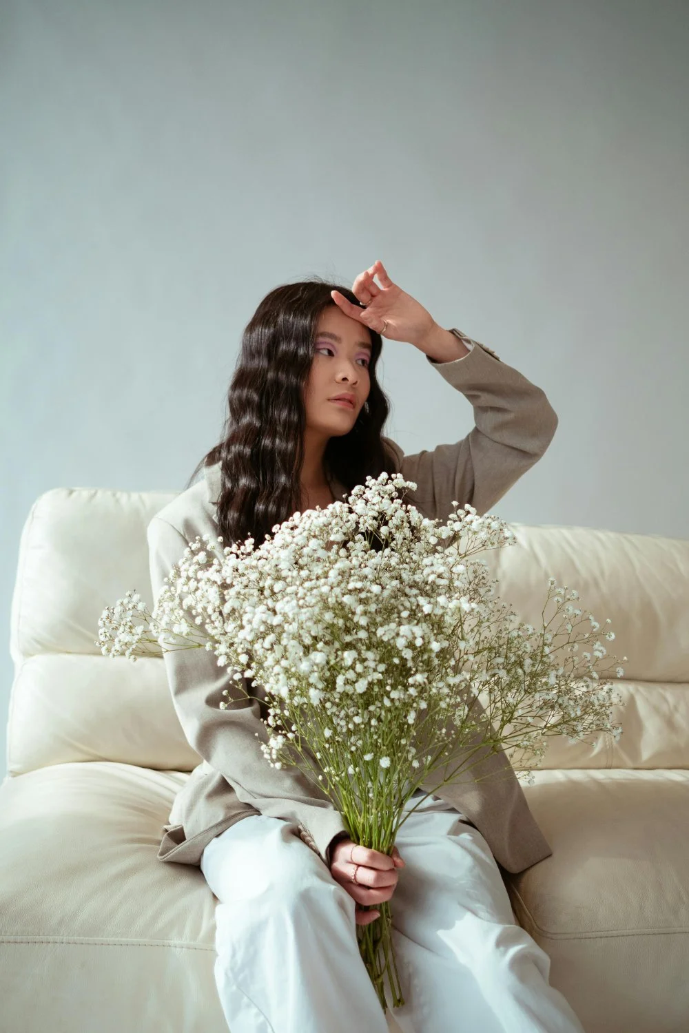Woman with long wavy hair sitting on a cream sofa, holding a bouquet of white baby's breath flowers, dressed in a beige blazer and white pants, with her right hand raised to her forehead, looking contemplative.