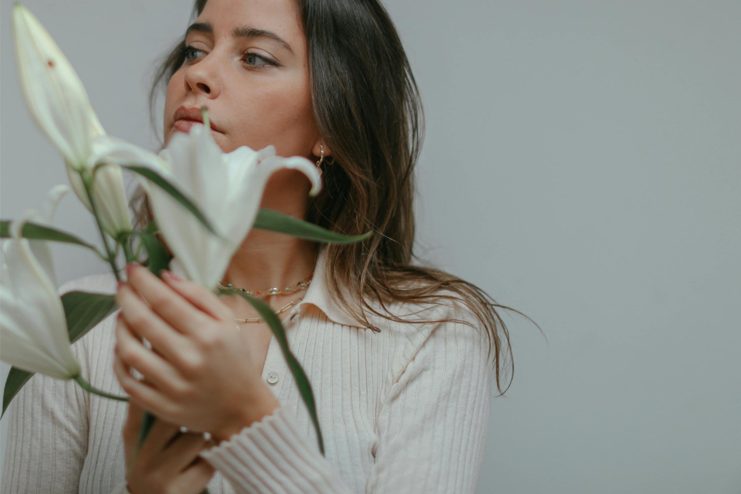 A woman with long brown hair holding a bouquet of white lilies, partially covering her face, against a plain grey background.