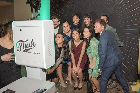 Two women standing indoors, reading papers, one holding a microphone, in front of a wooden wall.