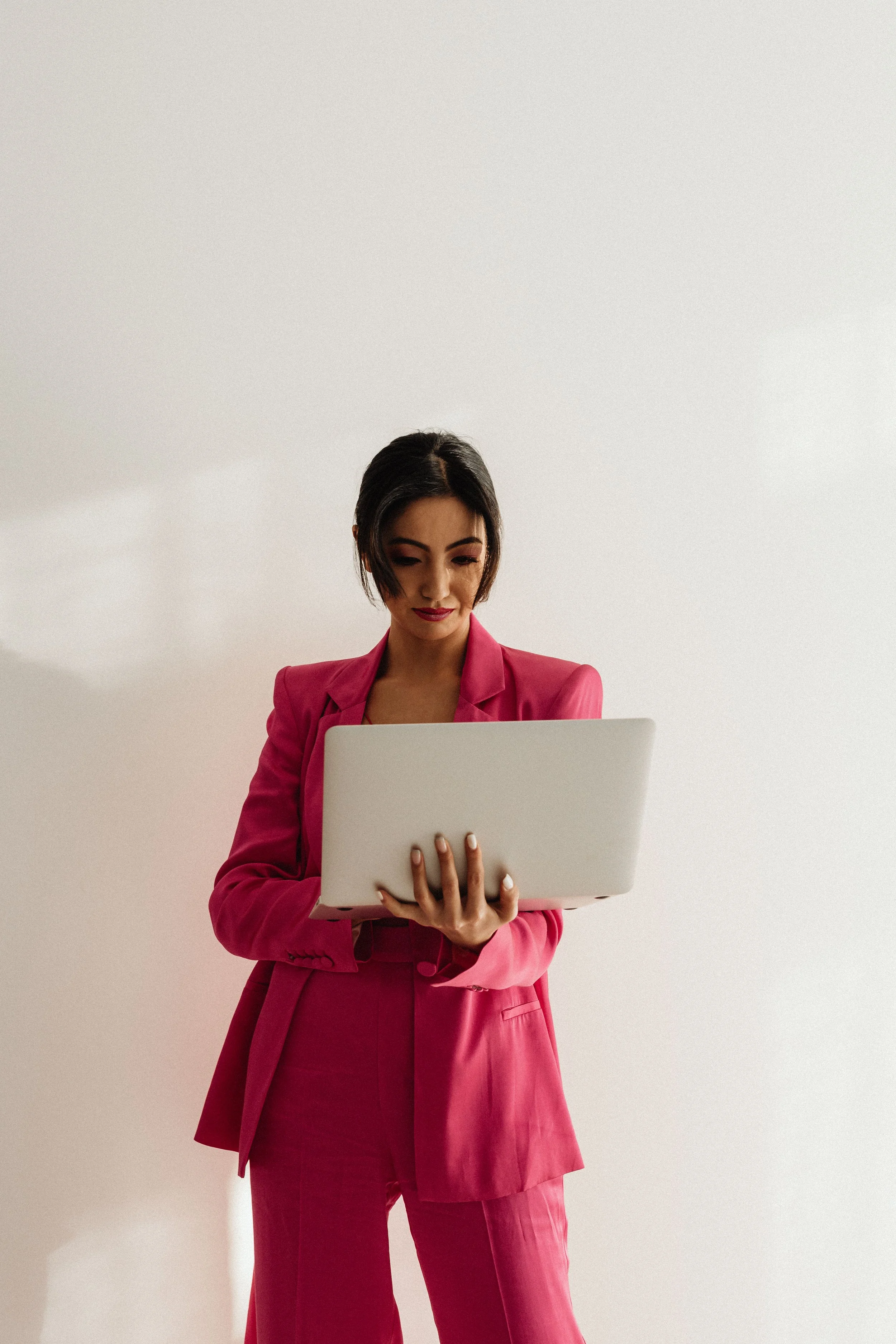 A woman in a pink suit holding a laptop and looking down at it, standing against a plain white wall.