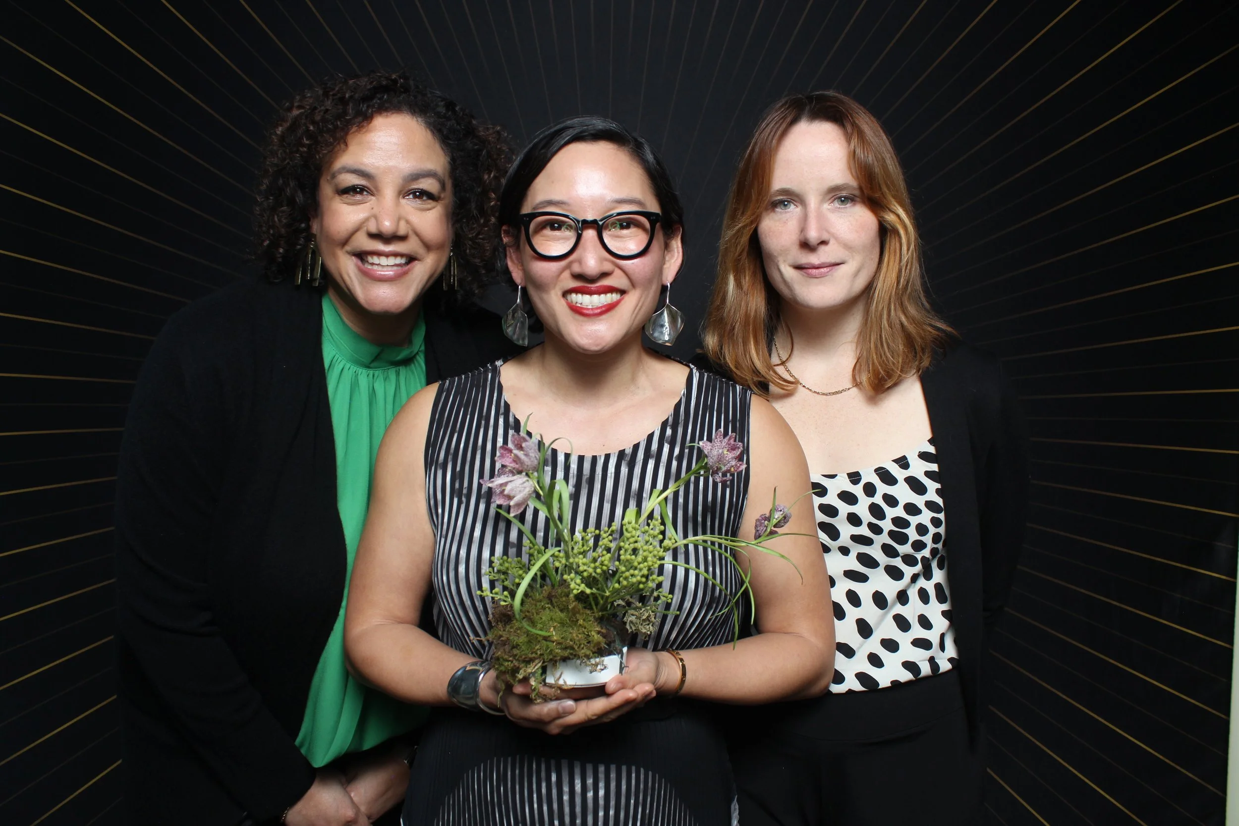 A woman in a pink suit holding a laptop and looking down at it, standing against a plain white wall.