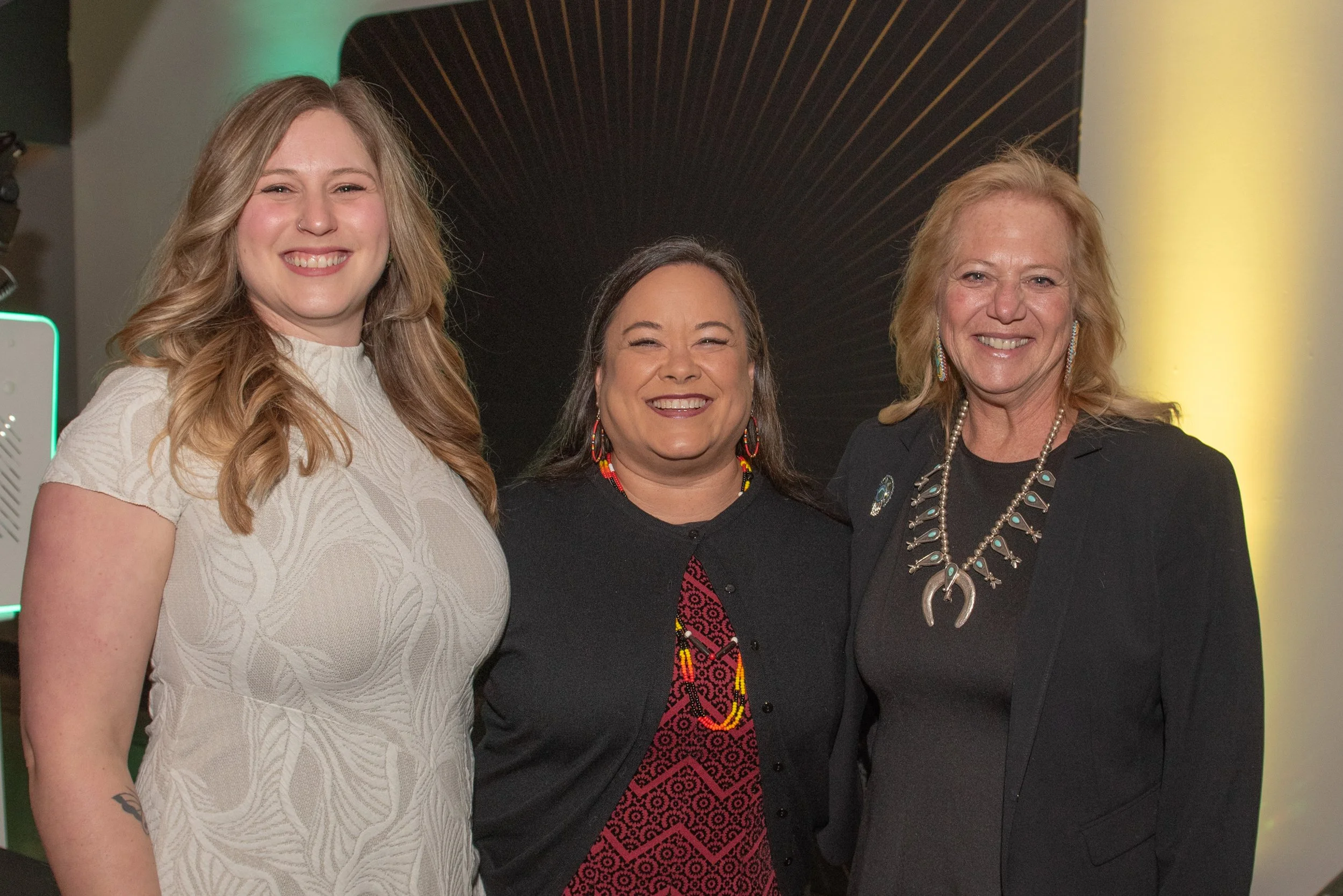 Two women standing indoors, reading papers, one holding a microphone, in front of a wooden wall.