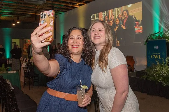 A person sitting on a red couch, holding a smartphone displaying an the Center for Women's Leadership Instagram profile, with part of their floral-patterned blouse and jeans visible.