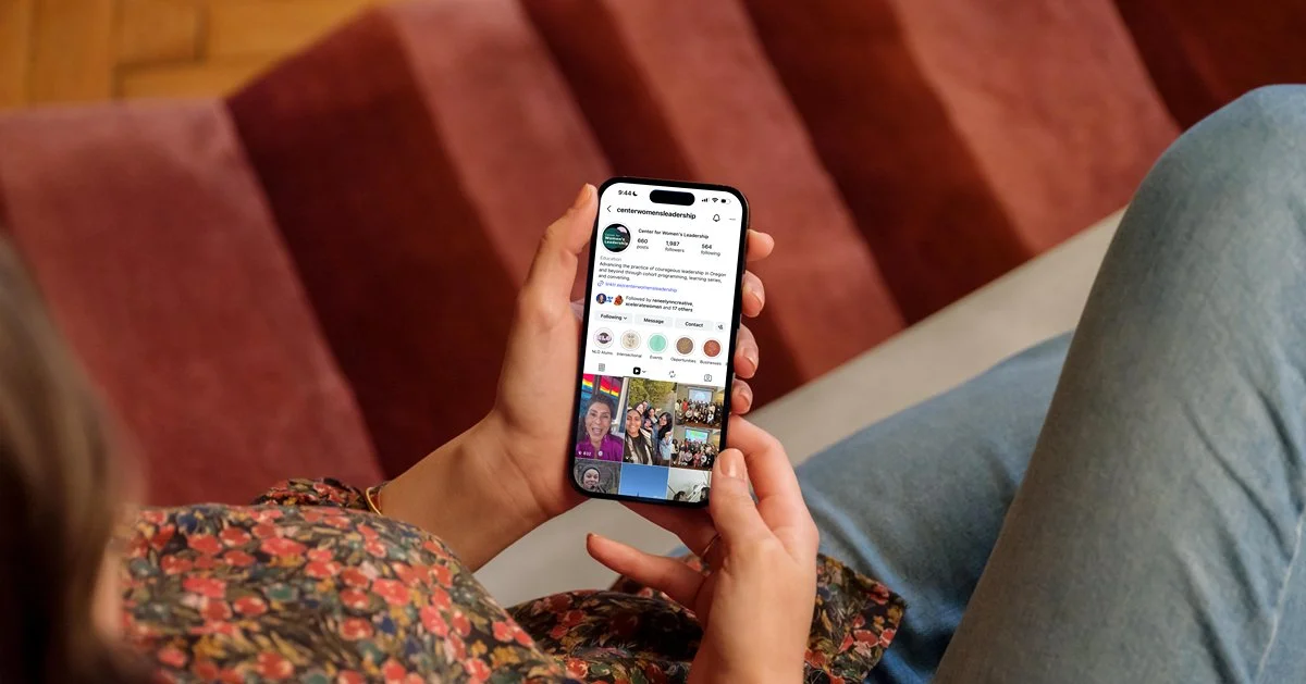 A person sitting on a red couch, holding a smartphone displaying an the Center for Women's Leadership Instagram profile, with part of their floral-patterned blouse and jeans visible.