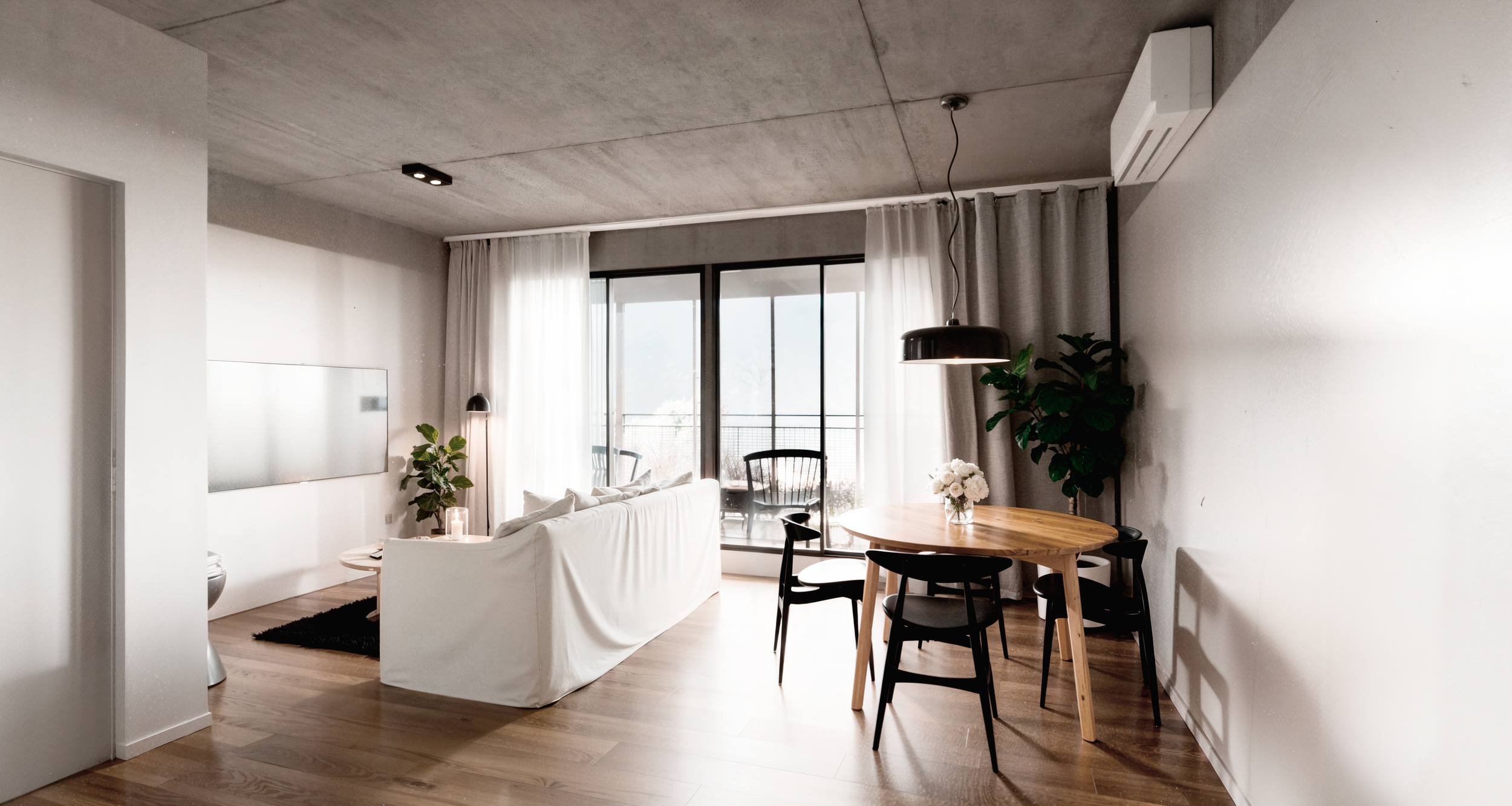 Modern living room with white sofa, round wooden dining table with black chairs, large window with sheer curtains, and indoor plants.