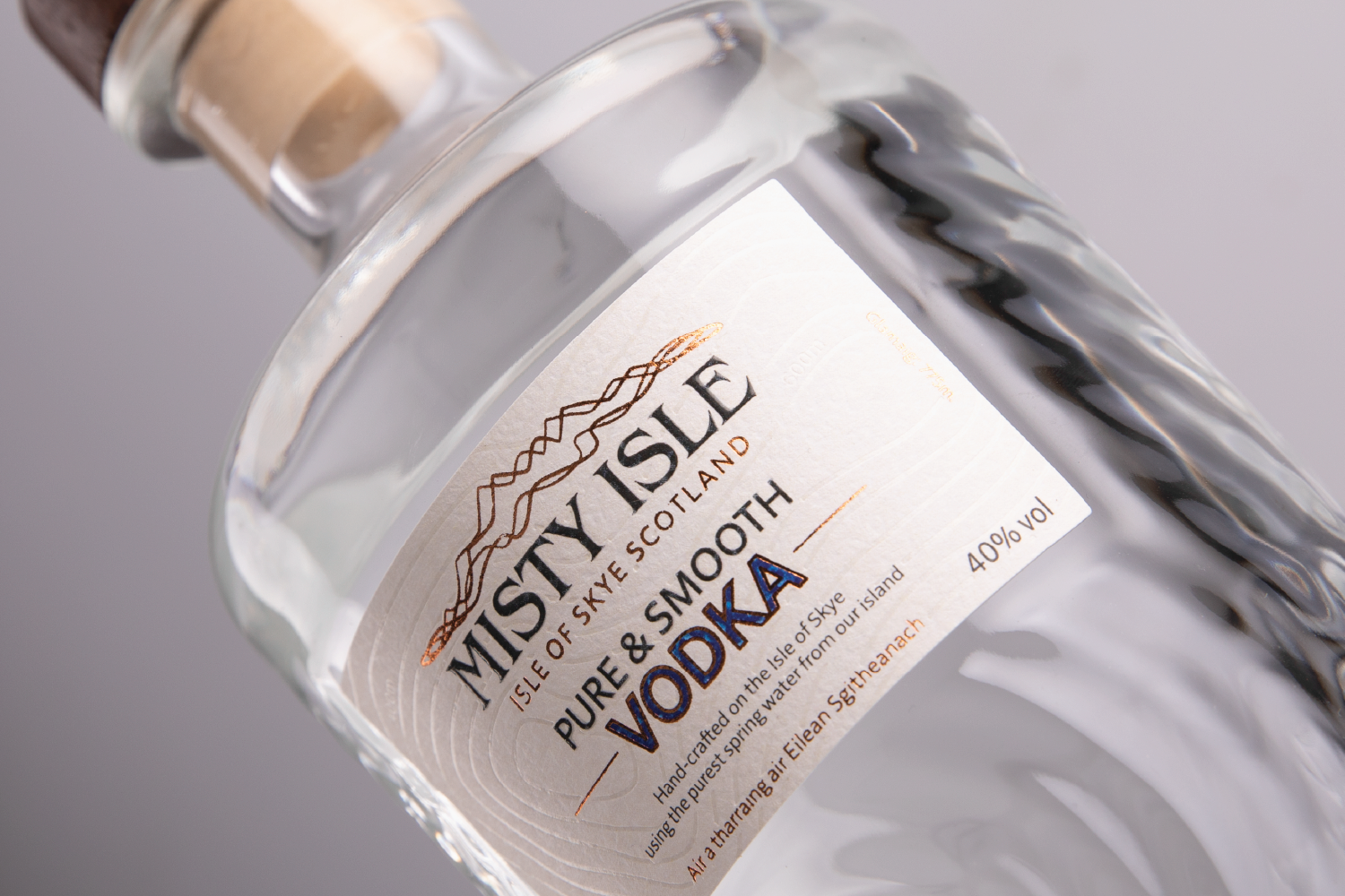 Close-up of a bottle of Misty Isle vodka, with a textured glass body and a label indicating Scottish origin, 40% alcohol by volume, featuring mountain graphics and orange accent text.