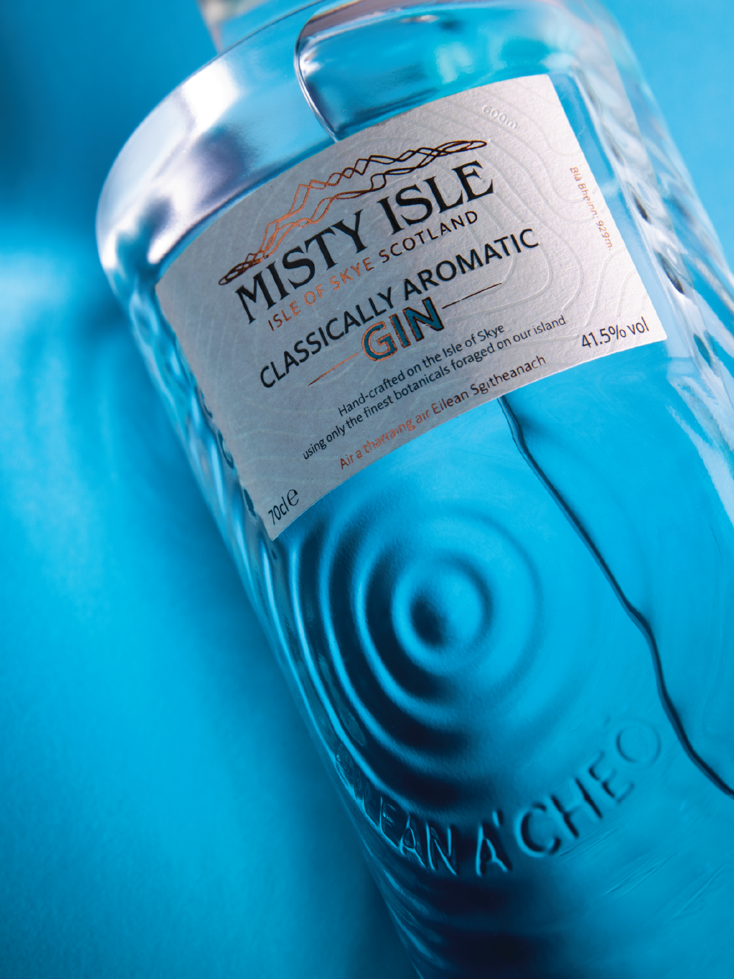 A clear glass bottle of Misty Isle gin with a blue background. The label indicates it is an aromatic gin from the Isle of Skye, Scotland, with 41.5% alcohol by volume.