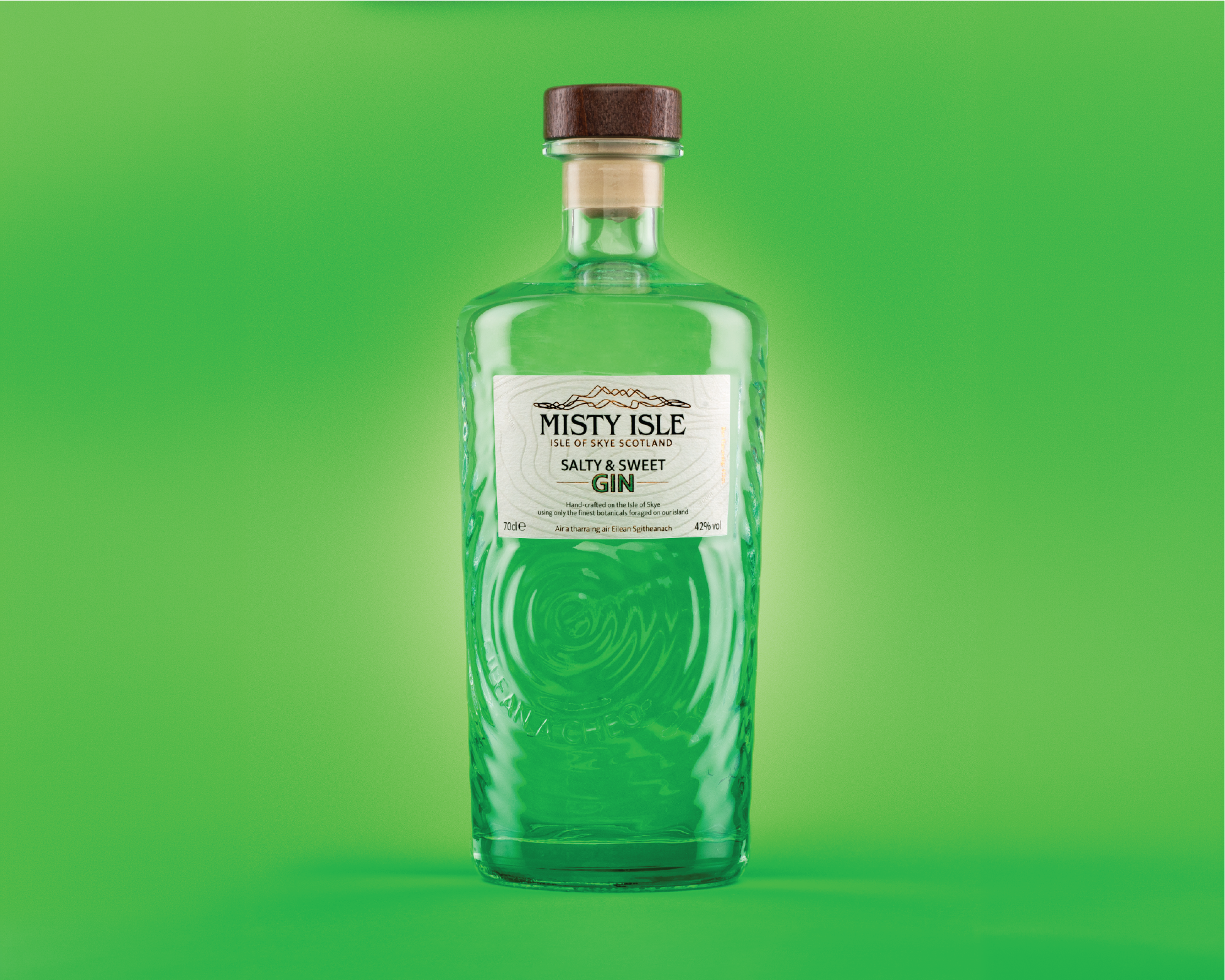 A clear glass bottle with a wooden cap labeled 'Misty Isle Salty & Sweet Gin', containing green-colored liquid, against a matching green background.