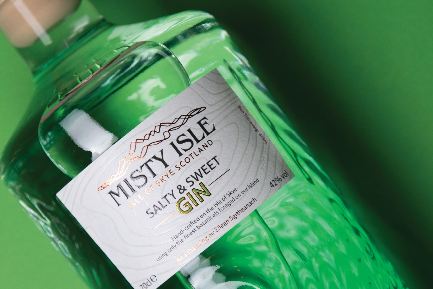 Close-up of a bottle of Misty Isle Salty & Sweet Gin with a white label and green liquid, against a green background.
