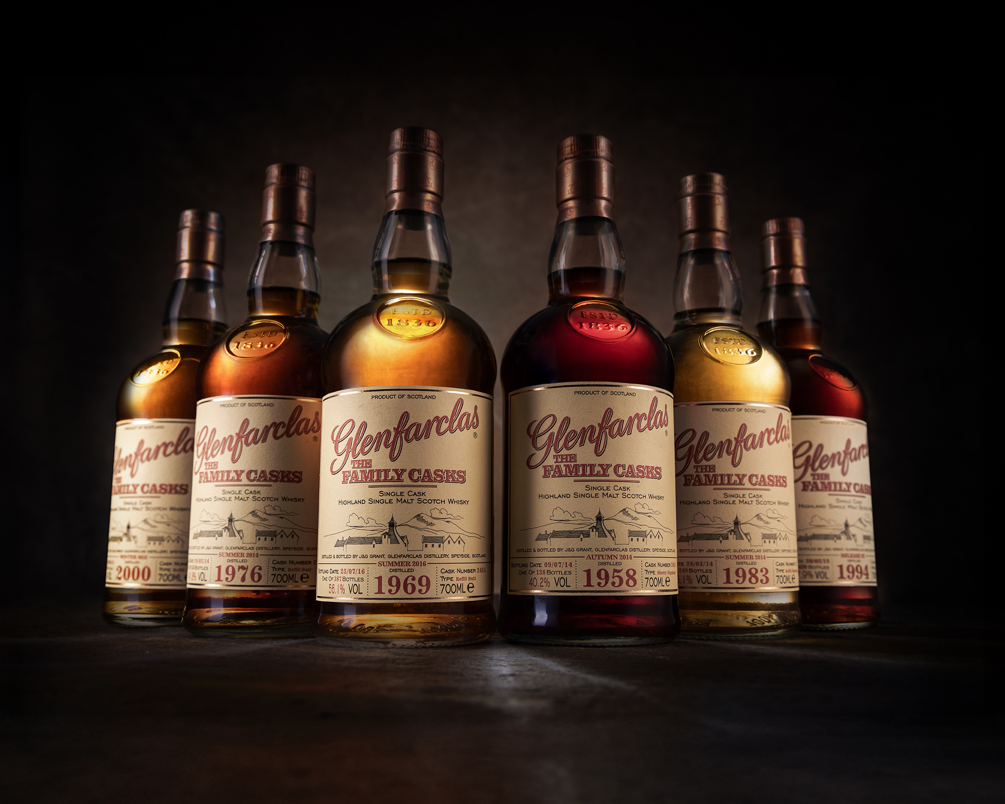 Six bottles of Glenfarclas single malt Scotch whisky arranged in a semi-circle on a dark surface, with a dark background. The labels indicate different cask years between 1969 and 2000.