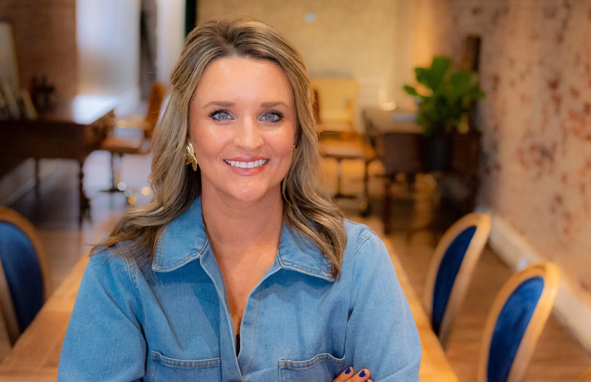 Jessica Sanford, a smiling woman with blue eyes and brown hair, wearing a denim shirt, sitting in a her office in Alexadner City, AL with a long wooden table and blue chairs, and a potted plant in the background.