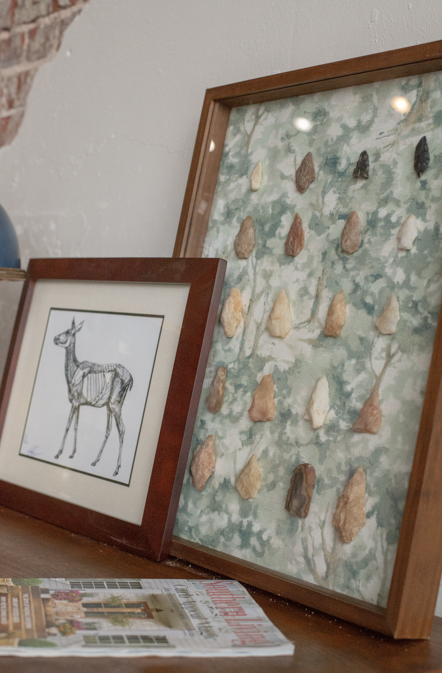 A framed drawing of a deer and a framed display of various arrowheads with a leaf-patterned background, placed on a surface in front of a brick wall.