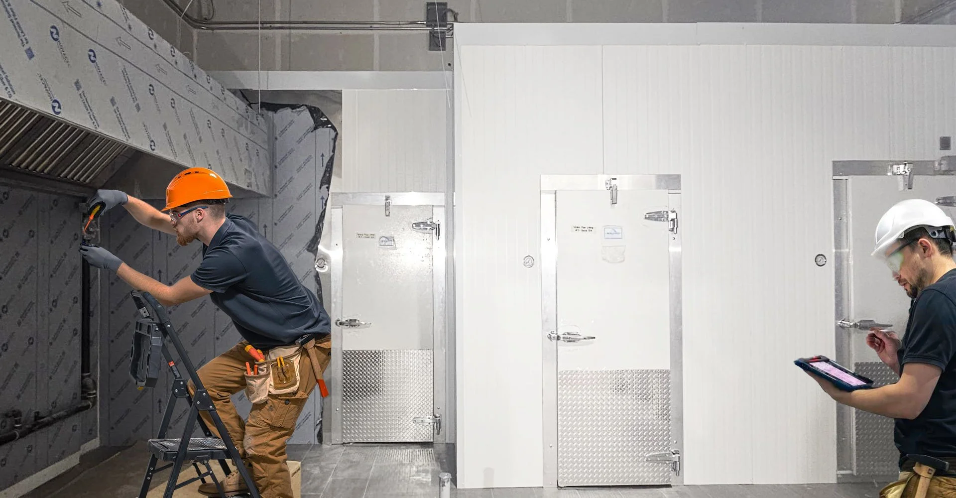 Two construction workers with hard hats and tools working inside an industrial facility. One is on a ladder inspecting or repairing a panel, while the other is holding a tablet and taking notes or reviewing information.