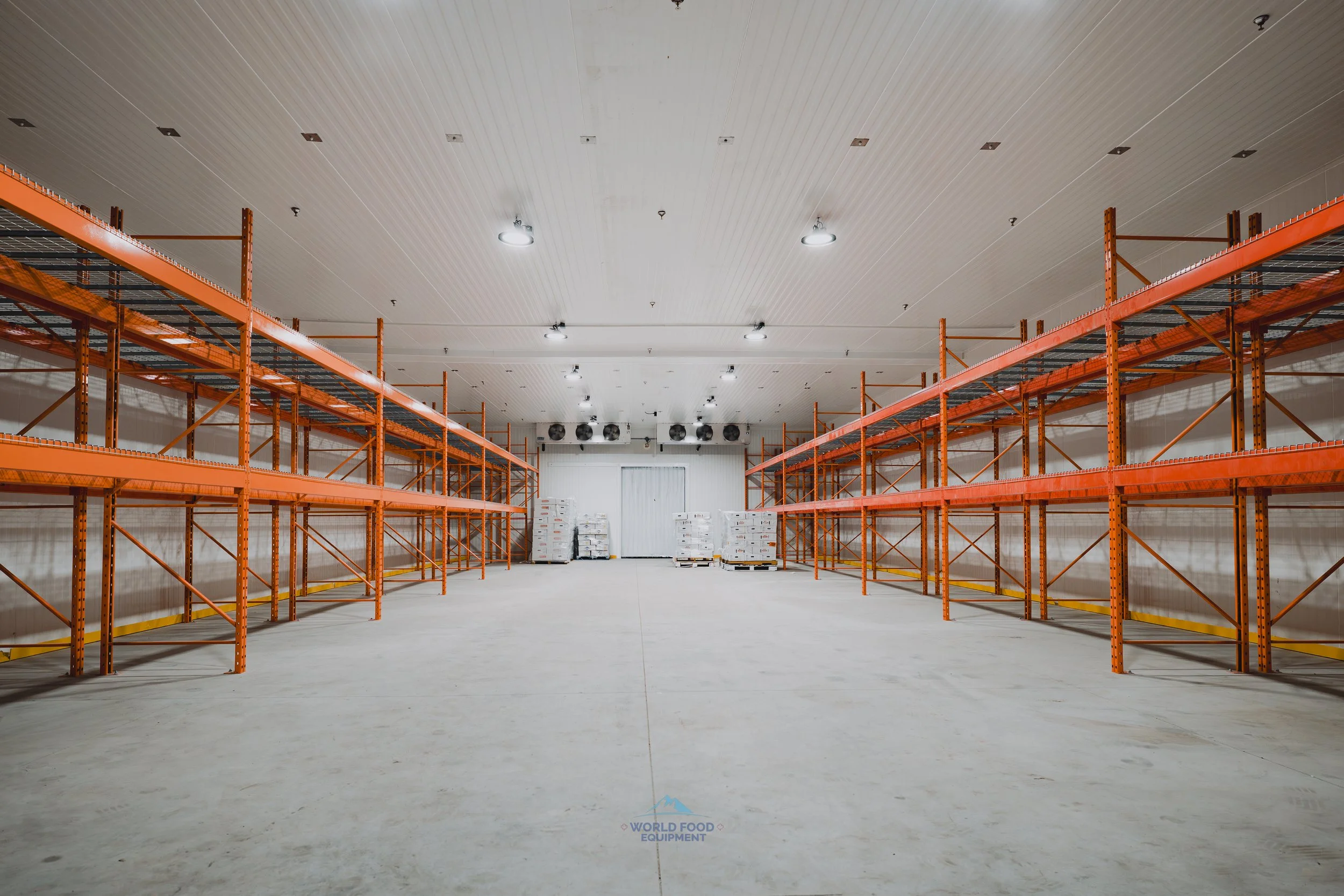Empty warehouse with orange metal shelving units on both sides and pallets of goods in the back.