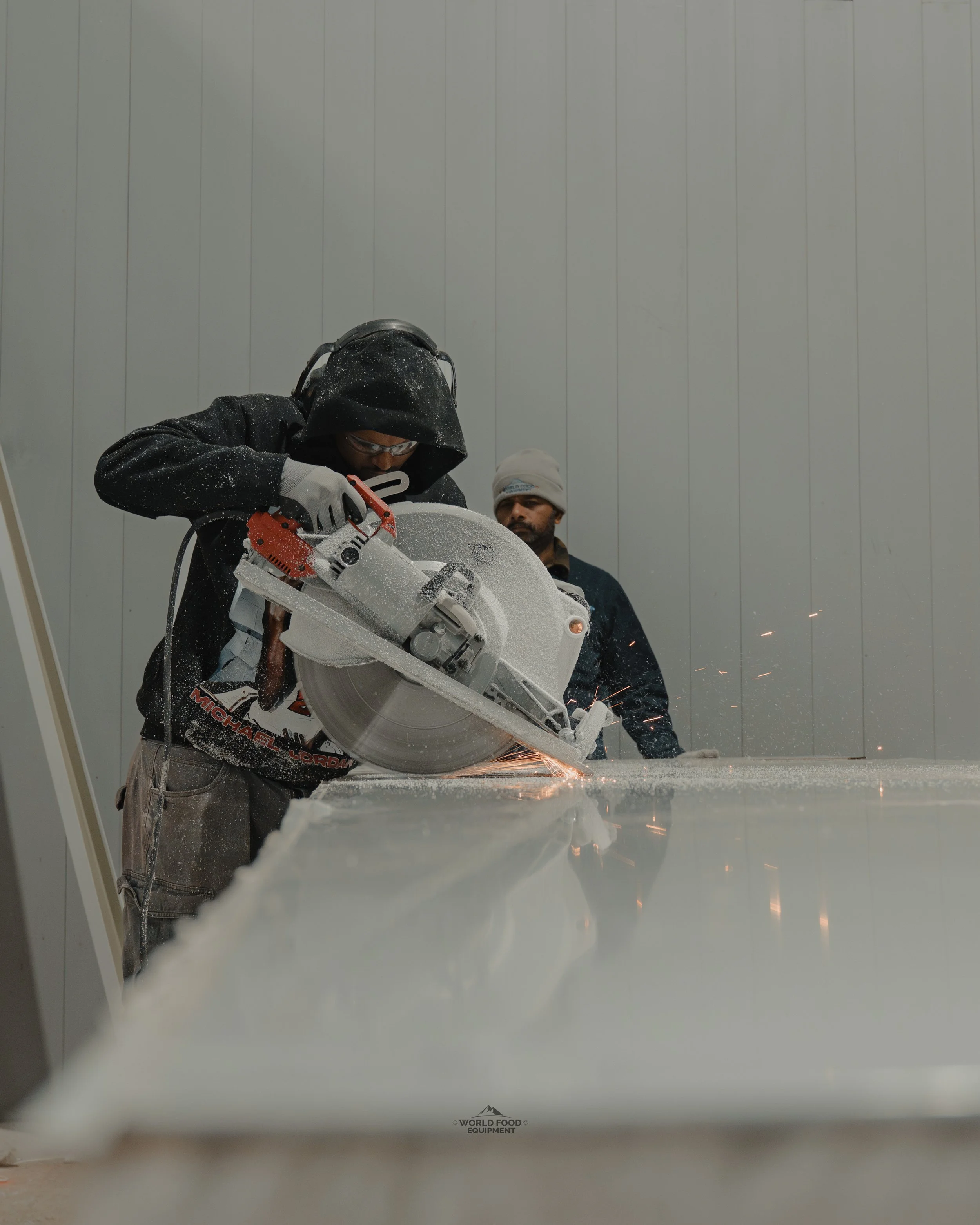 A person wearing safety gear using a large circular saw to cut a material, with sparks flying, and another person observing in the background.