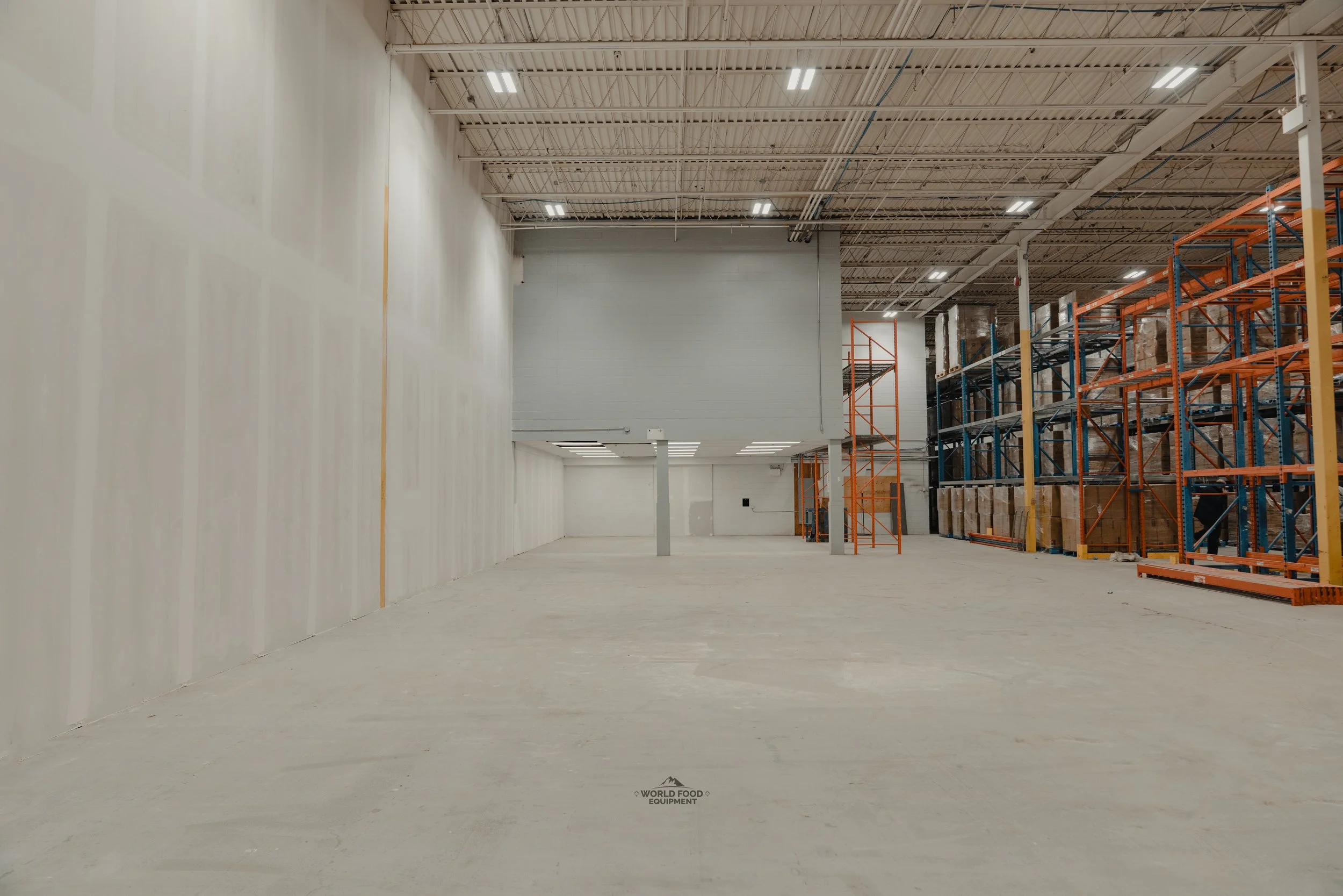 Empty warehouse with high ceilings, white walls, and metal shelving filled with boxes on the right side.