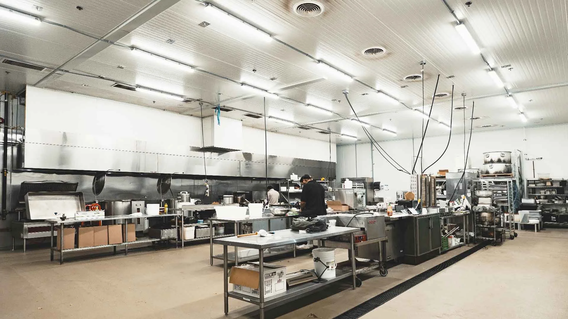 Commercial kitchen with stainless steel appliances, worktables, and workers preparing food, with overhead fluorescent lighting.