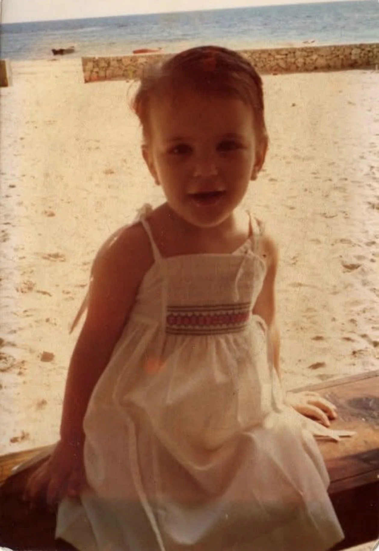 Niña de cabello corto y rubio en la playa, vistiendo un vestido blanco con detalles rojos, sentada junto a la arena y el mar al fondo.