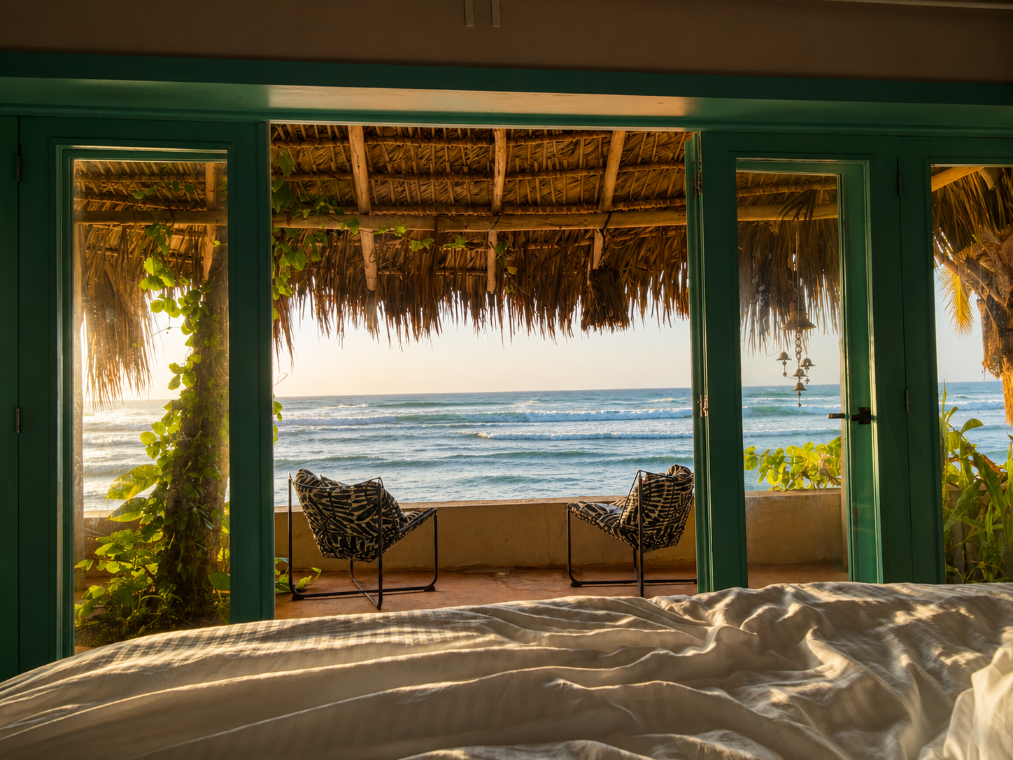 Vista desde una habitación de playa con puertas de vidrio que muestran una terraza con sillas y vista al mar con olas y cielo al atardecer.
