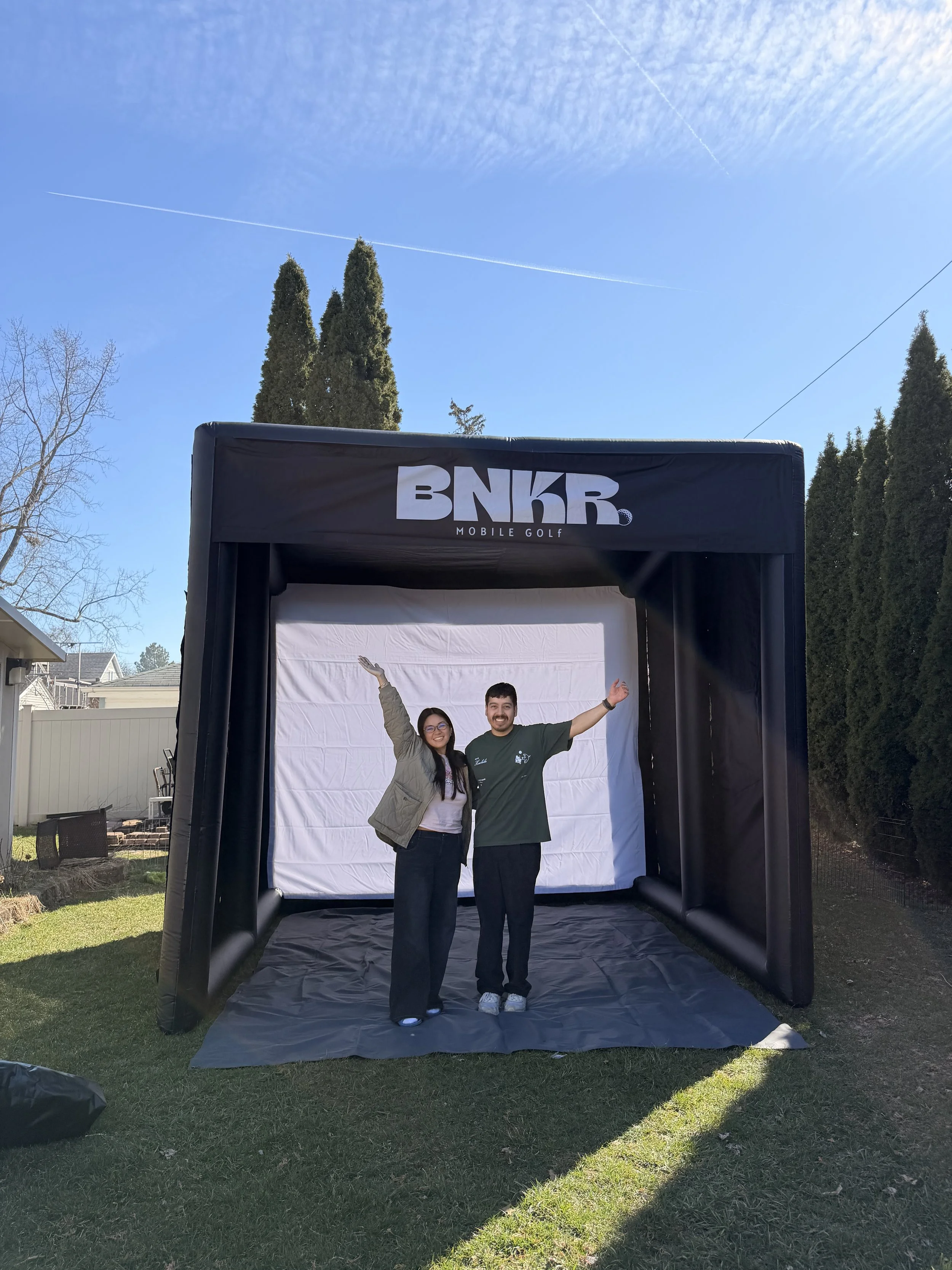 Two people, a woman and a man, stand inside a portable inflatable screen setup labeled 'BNKR Mobile Golf'. They are smiling and waving outdoors on a sunny day. The background features a clear blue sky, tall trees, and a residential neighborhood.