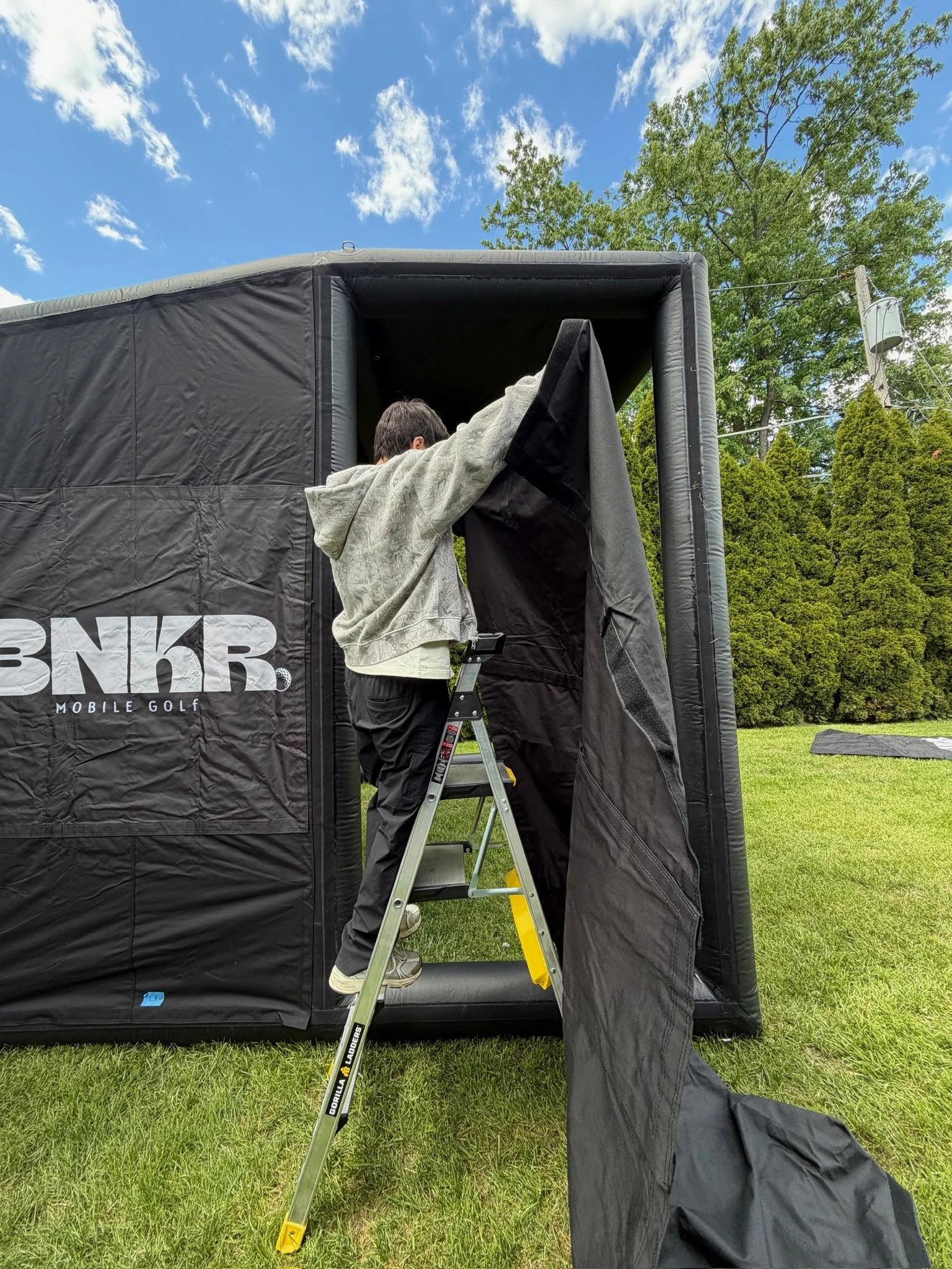 A person climbing a ladder into a black portable golf net outdoor on a grassy area under a blue sky with clouds and trees.