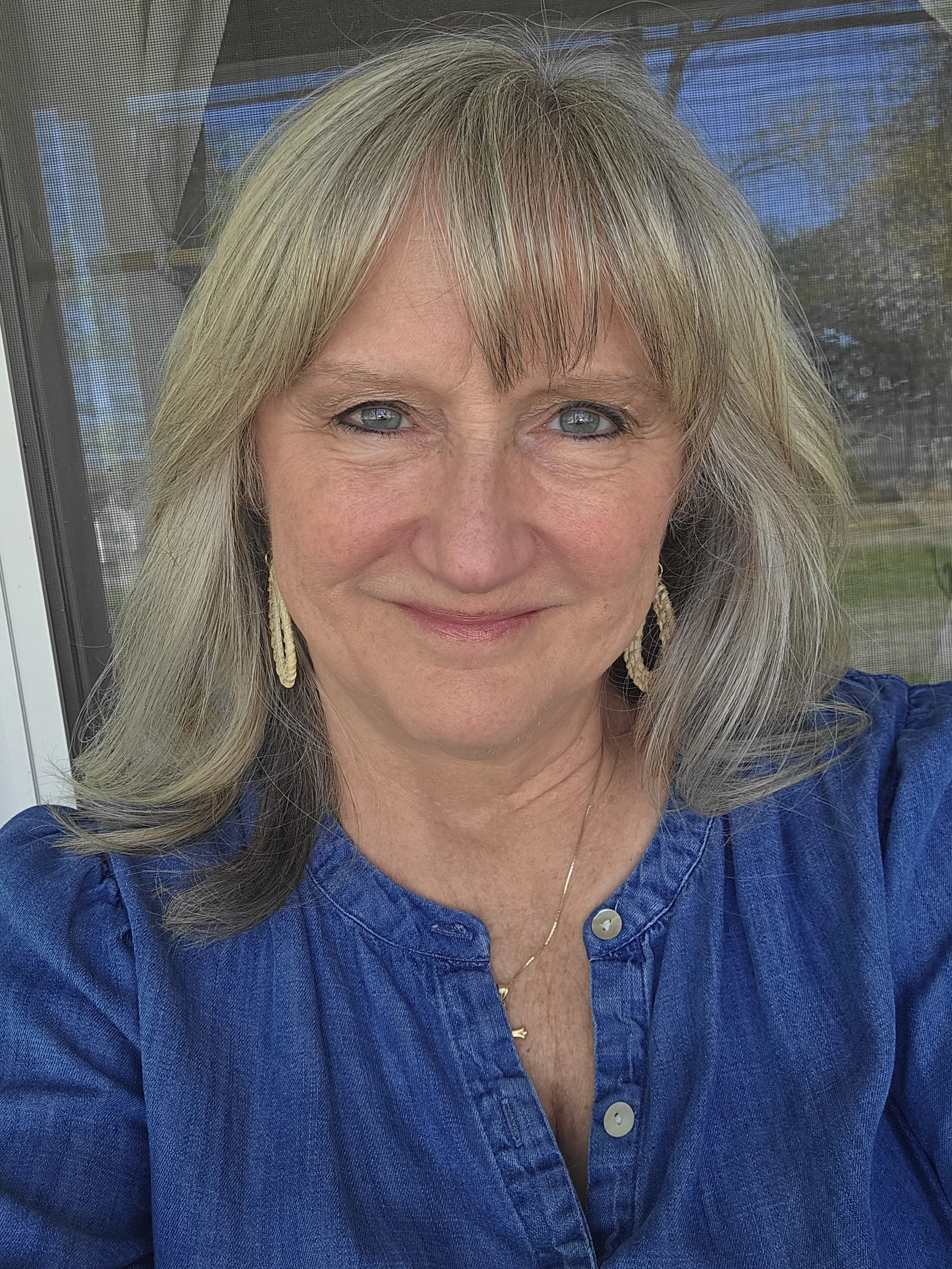 A woman with blond hair, blue eyes, and wearing a blue top and gold earrings, smiling outdoors with a screened porch and trees in the background.