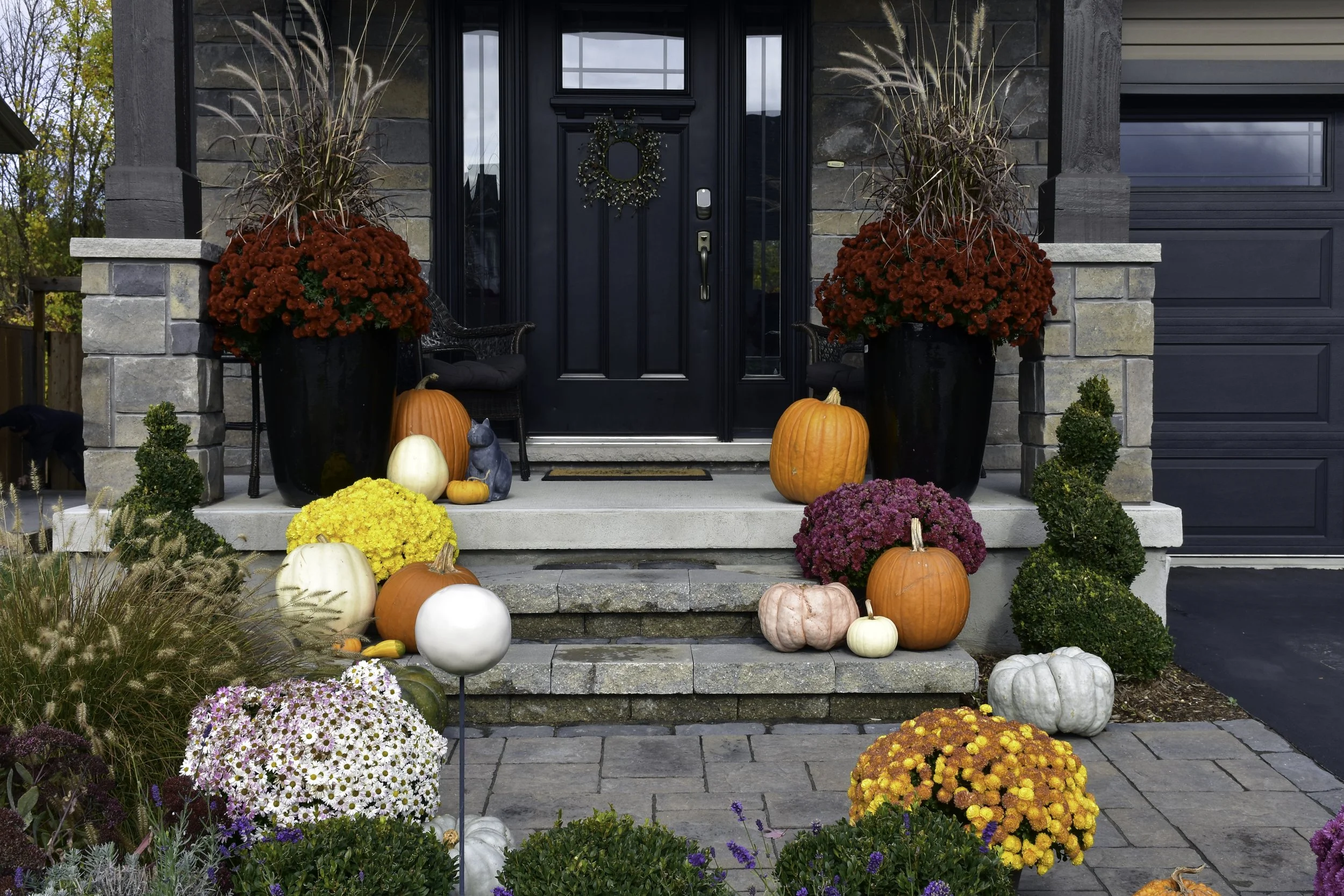 A front porch decorated for fall with pumpkins, gourds, and colorful chrysanthemums, flanked by two large black planters with red flowering plants, and small bushes along the steps.