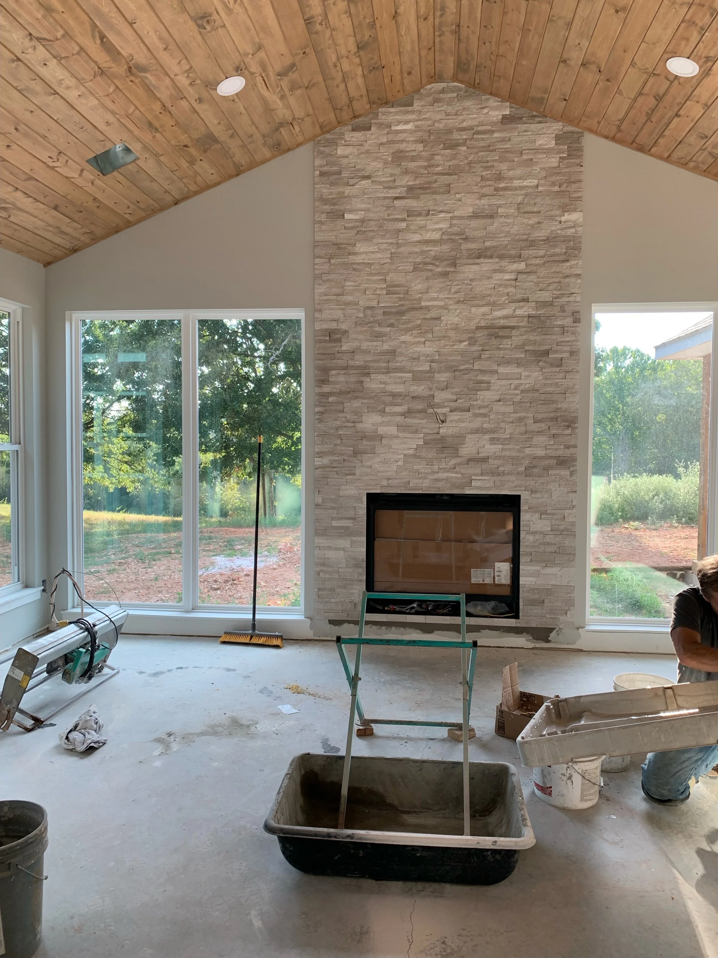 Living room under construction with a stone fireplace, large windows, a vaulted wooden ceiling, construction tools and materials, and a worker on the right