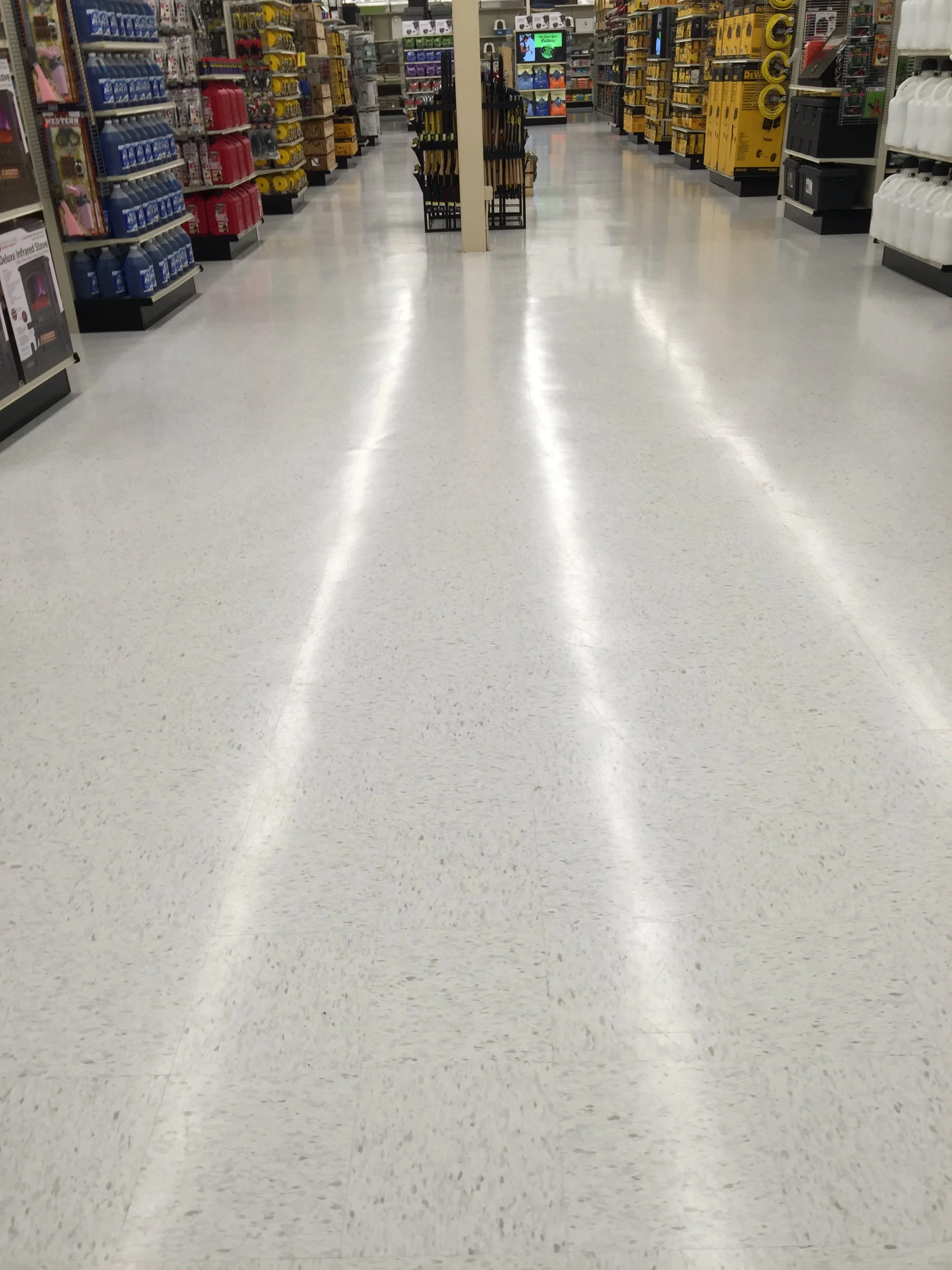 Empty aisle in a hardware store with shelves stocked with various tools and supplies on both sides. Floors done by White Knight Janitorial.