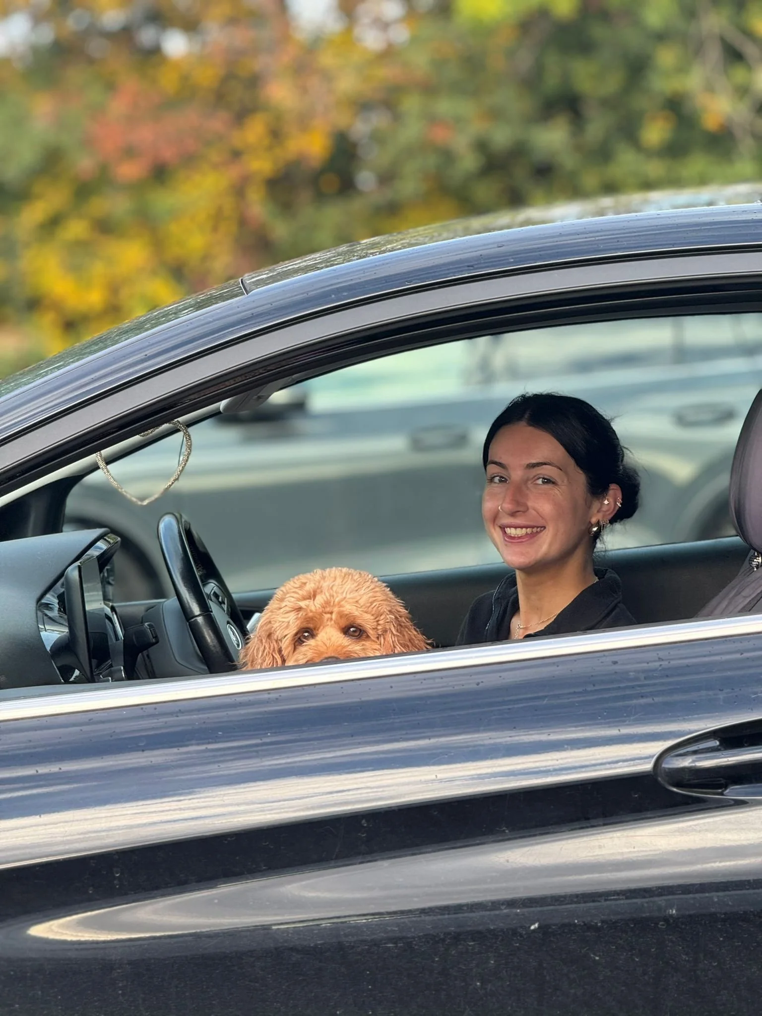 A woman with dark hair and a dog with curly fur inside a car, looking through the open window. The woman is smiling and the background shows autumn foliage.