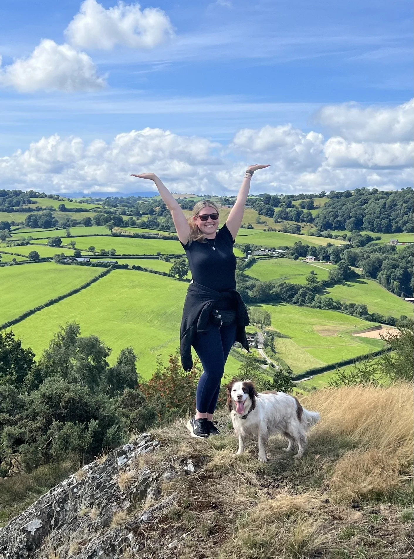 A woman with sunglasses and a black shirt smiling with her arms raised standing on a hill with a white and brown dog, overlooking green rolling fields and a blue sky with clouds.