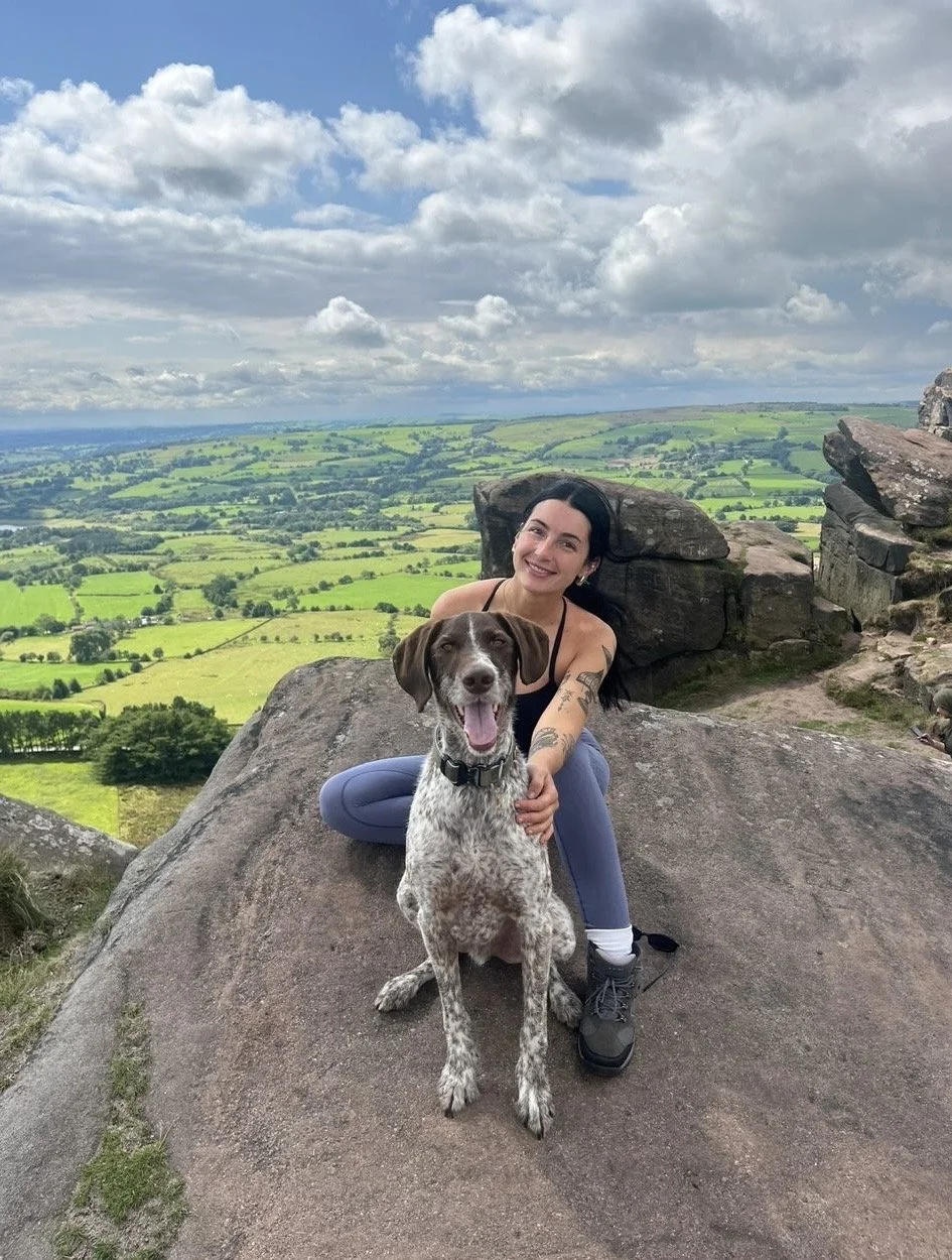 A woman with black hair and tattoos on her arm, smiling and sitting on a large rock, holding a happy dog with brown and white fur and floppy ears, on a hilltop with a scenic green valley and cloudy sky in the background.