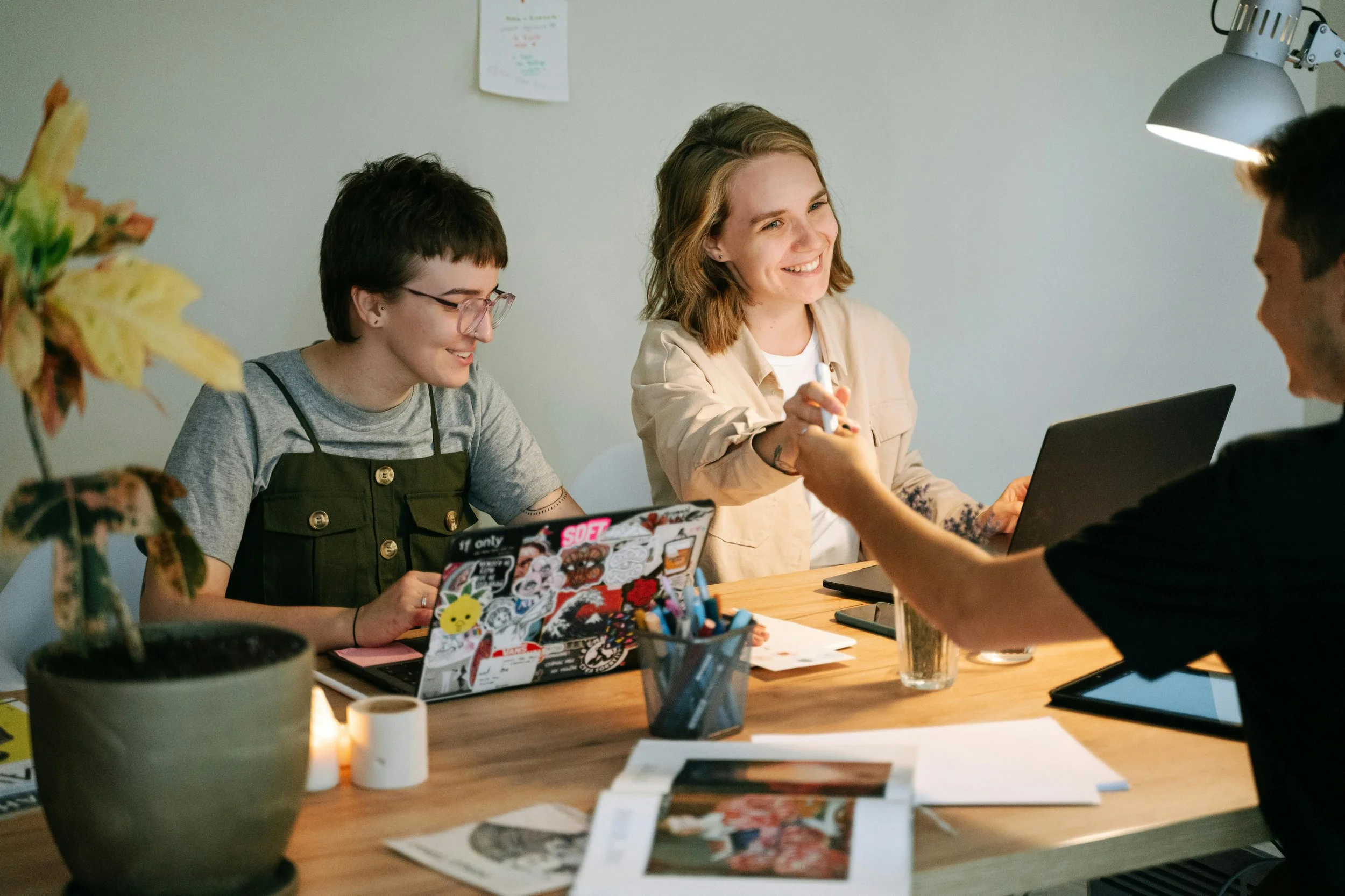 Four people sitting at a wooden table, engaging in a meeting or discussion. One person is smiling while another is reaching out to shake hands. The table has laptops, papers, and a container of pens, with a potted plant in the foreground.