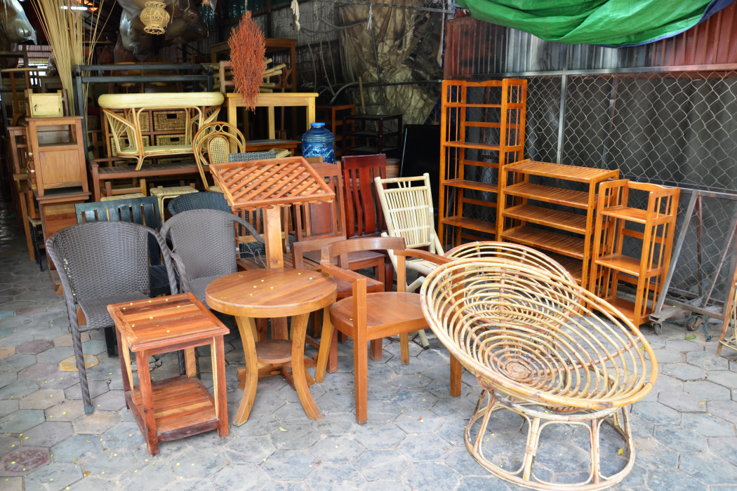 Collection of wooden and wicker furniture including chairs, tables, and shelves, arranged outdoors on a stone-paved surface.