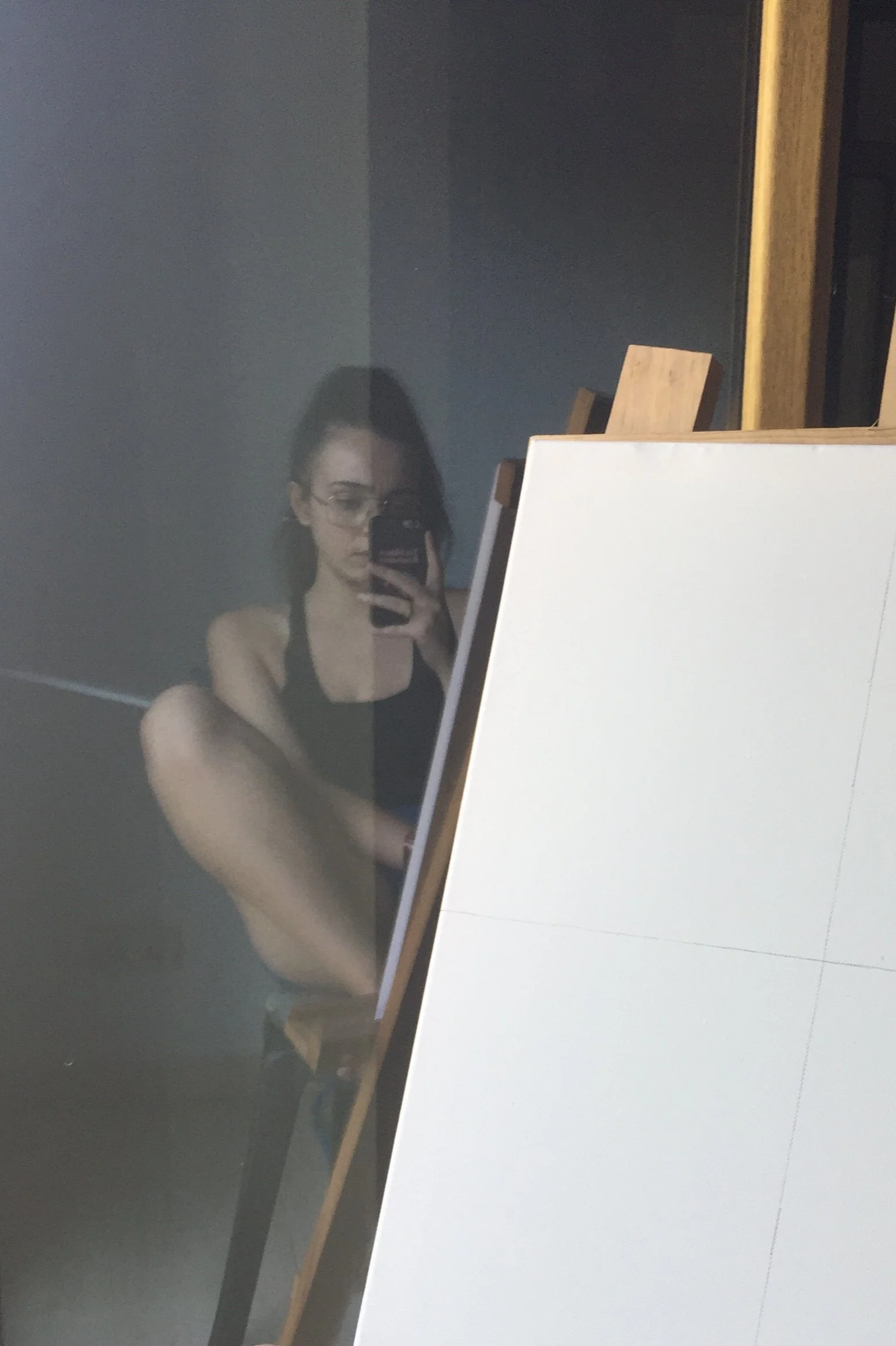 Reflejo de una mujer joven con gafas y ropa deportiva, tomando una selfie en una superficie reflectante, con una pared oscura y una silla de madera en el fondo.