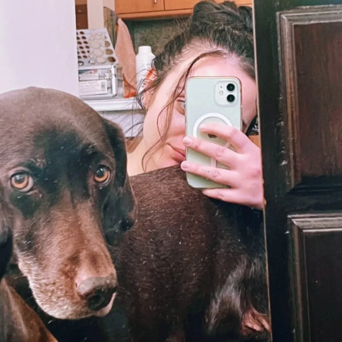 Una mujer toma una selfie en un espejo, sosteniendo un teléfono móvil con funda blanca, mientras un perro de pelo corto y color marrón oscuro se encuentra frente a ella, mirando hacía la cámara. El fondo tiene utensilios de cocina y una estantería.