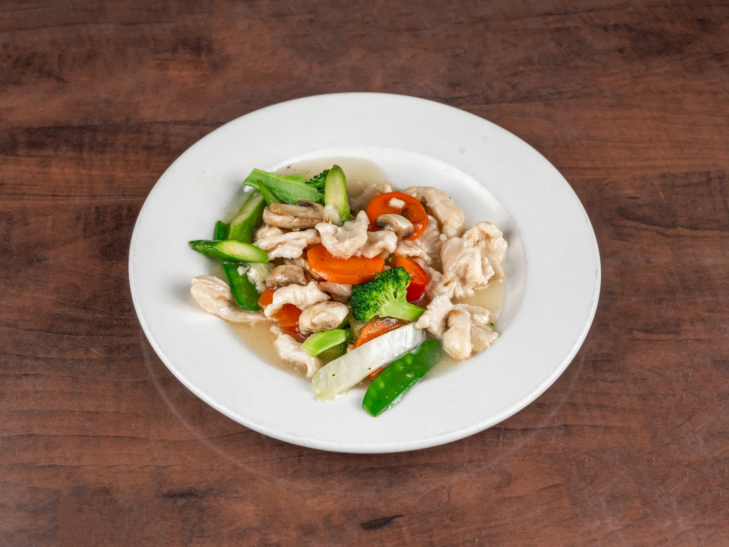 Stir-fried chicken with broccoli, carrots, snow peas, and mushrooms served on a white plate on a wooden table.