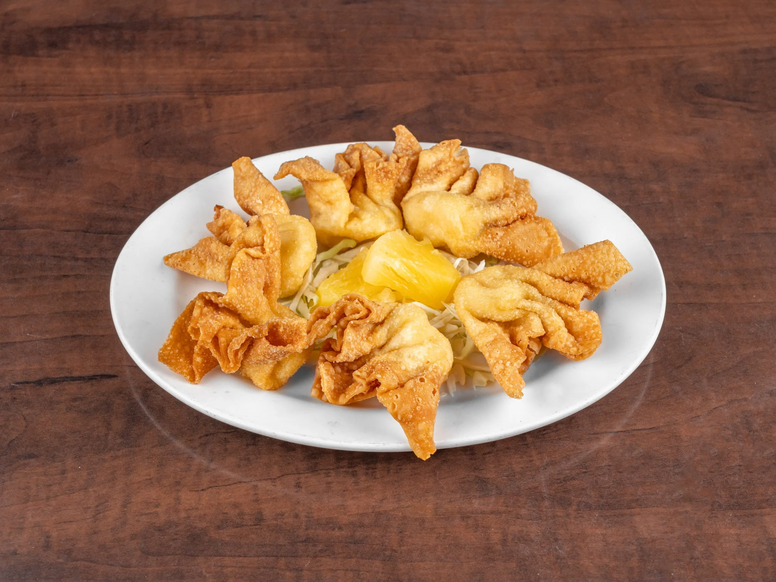 Fried wontons arranged in a circle on a white plate with a yellow pineapple chunk in the center, on a wooden table.
