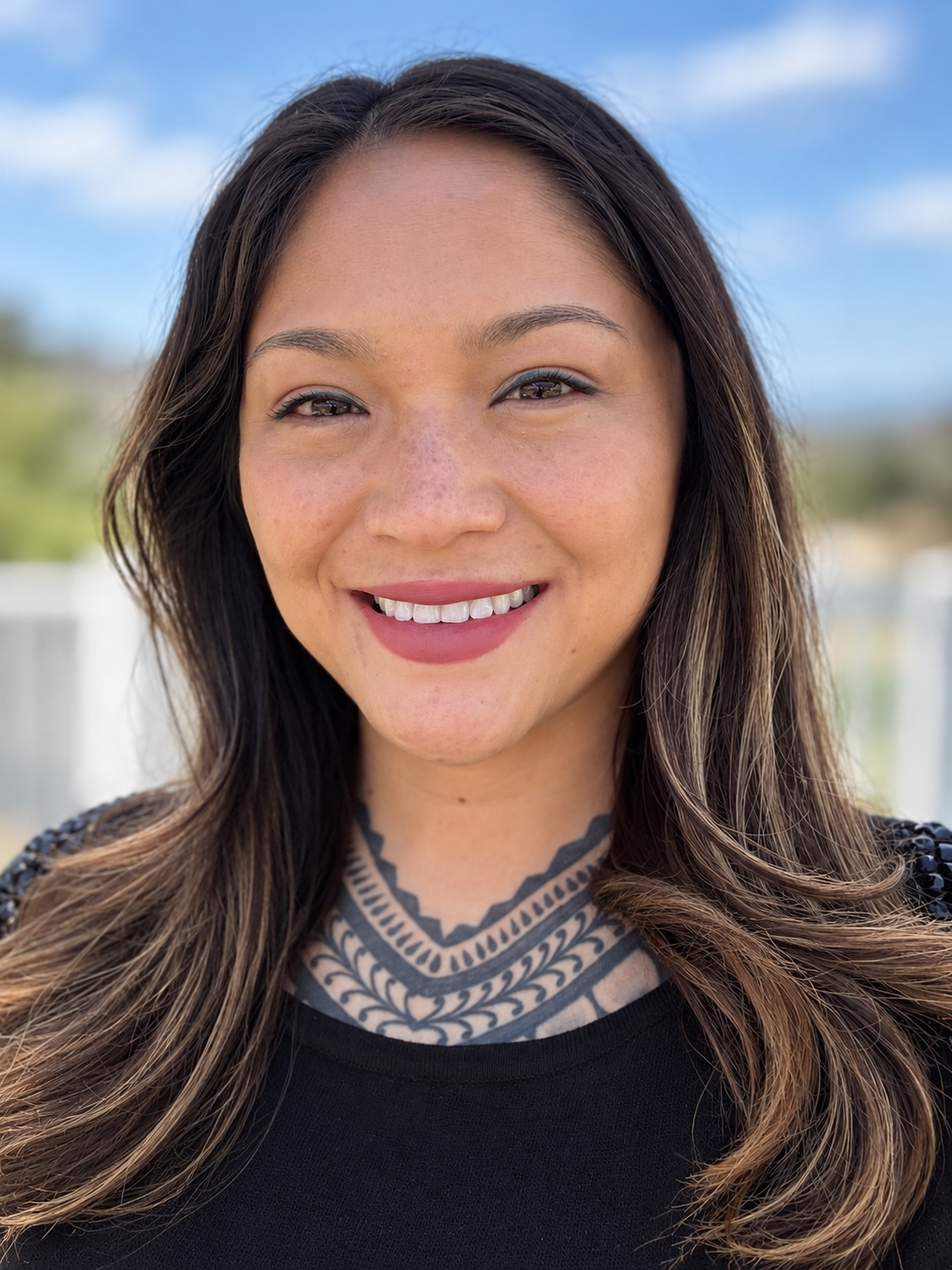 Close-up of a woman with long dark hair, tattoos on her neck, smiling outdoors with a blue sky and some blurred trees in the background. Marriage and family therapist, EMDR, psychotherapist, asian and Mexican descent, POC therapist.