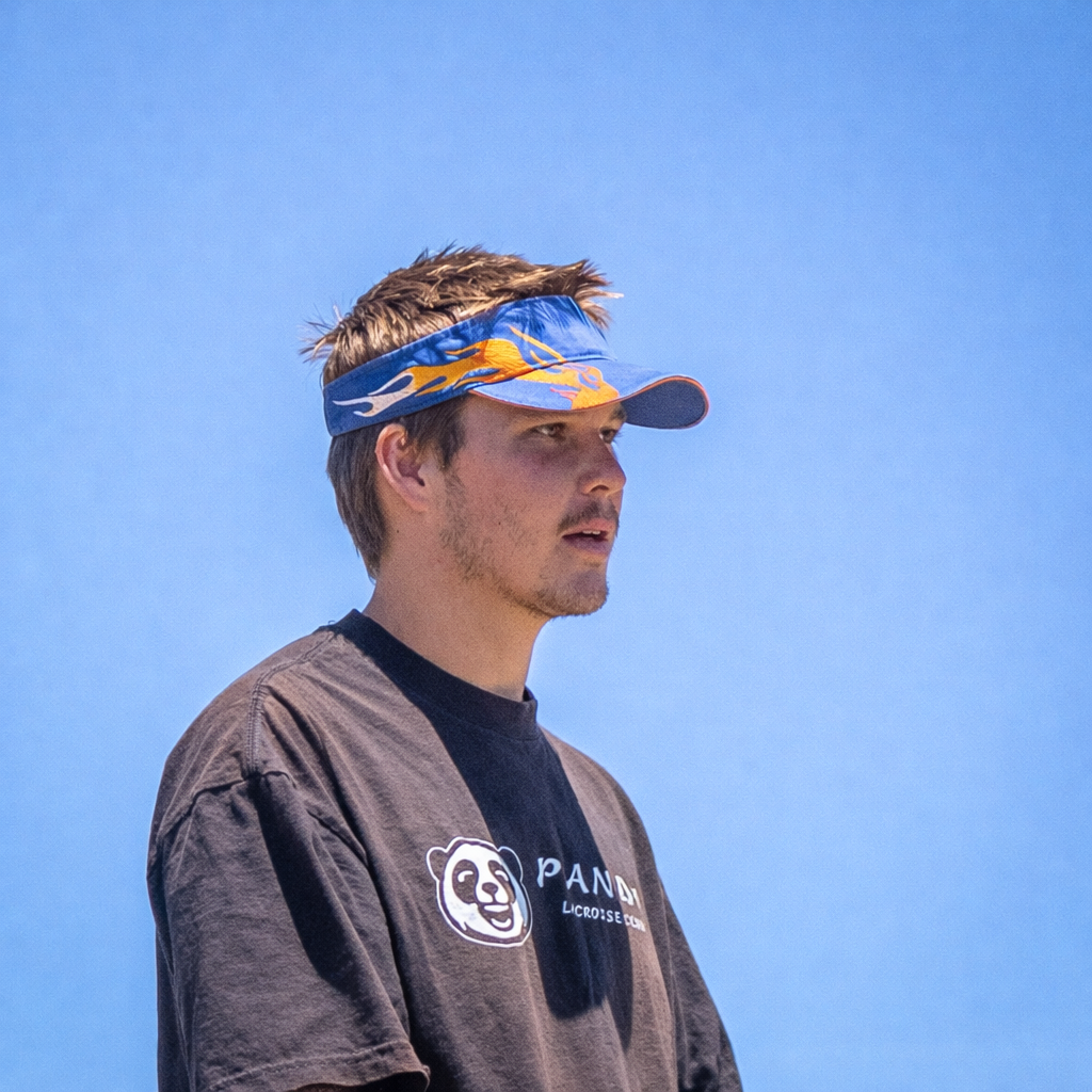 Young man wearing a blue visor with orange flames and a black T-shirt with a panda logo, standing outdoors against a clear blue sky.
