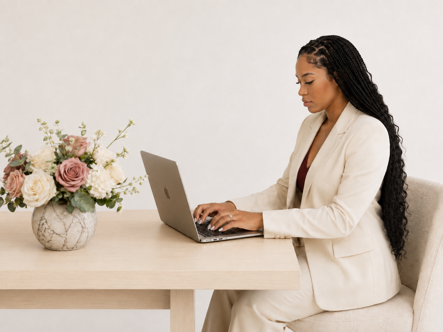 Anissia in a cream suit working on a laptop at a wooden desk with flowers.