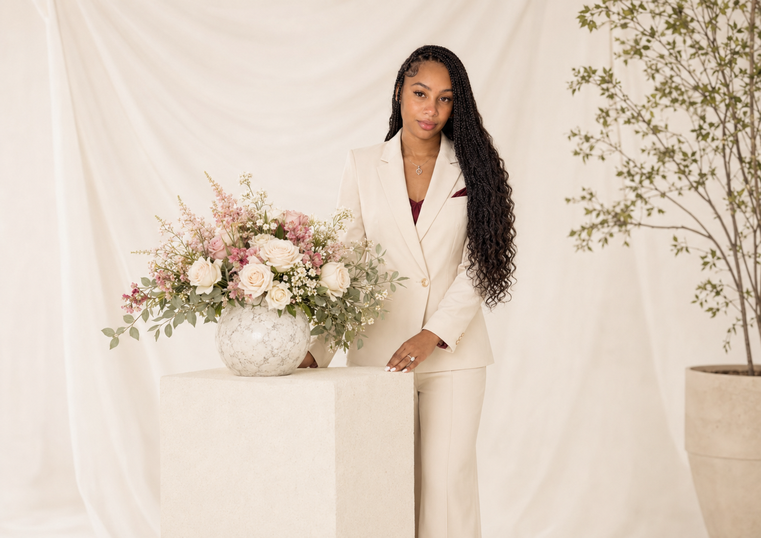 Anissia in a cream suit standing beside a white floral arrangement in a neutral studio.
