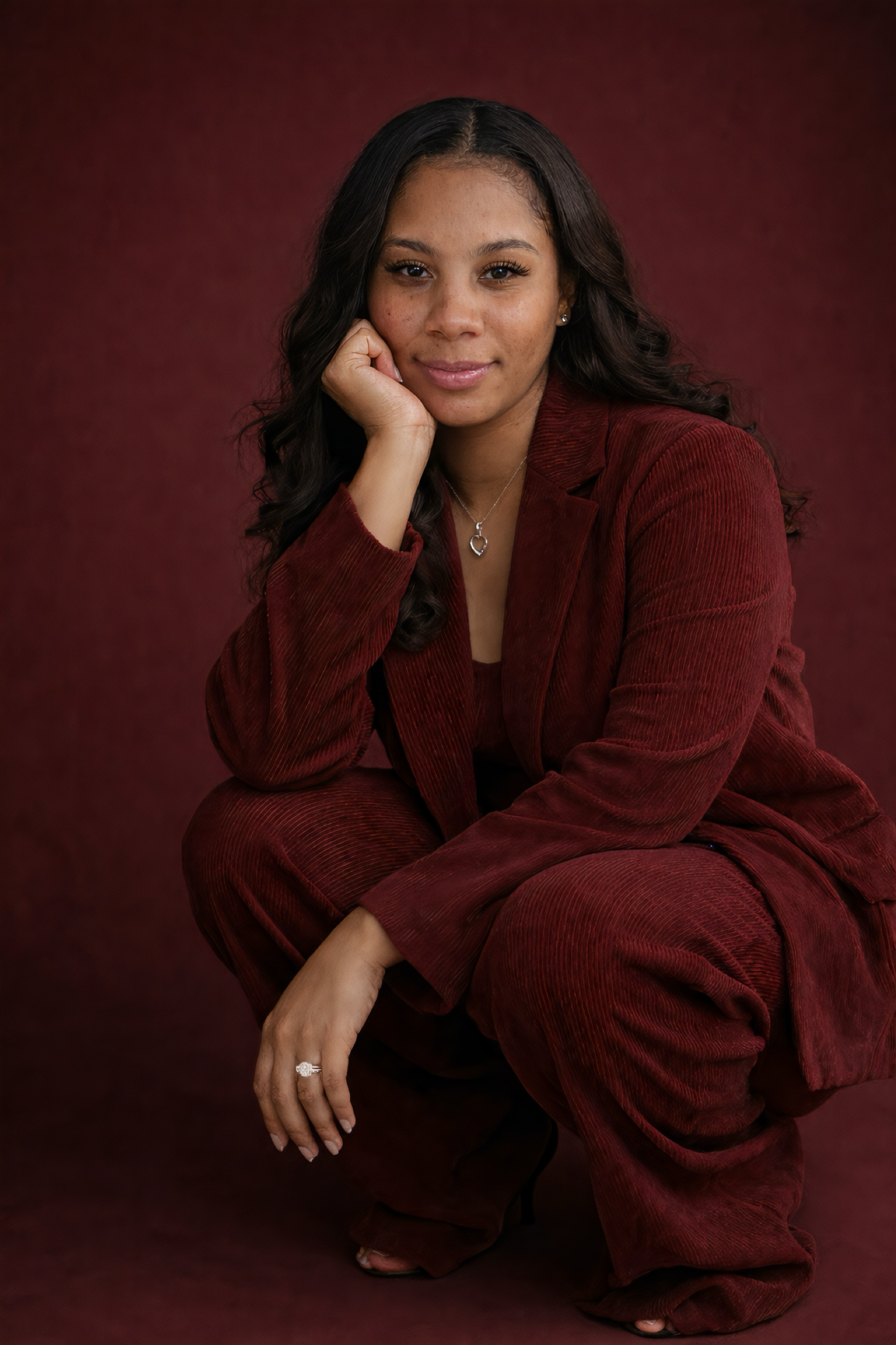 A woman with long dark hair, wearing a red velvet suit, crouches in a squatting position against a red background. She rests her chin on her left hand, smiling softly at the camera.